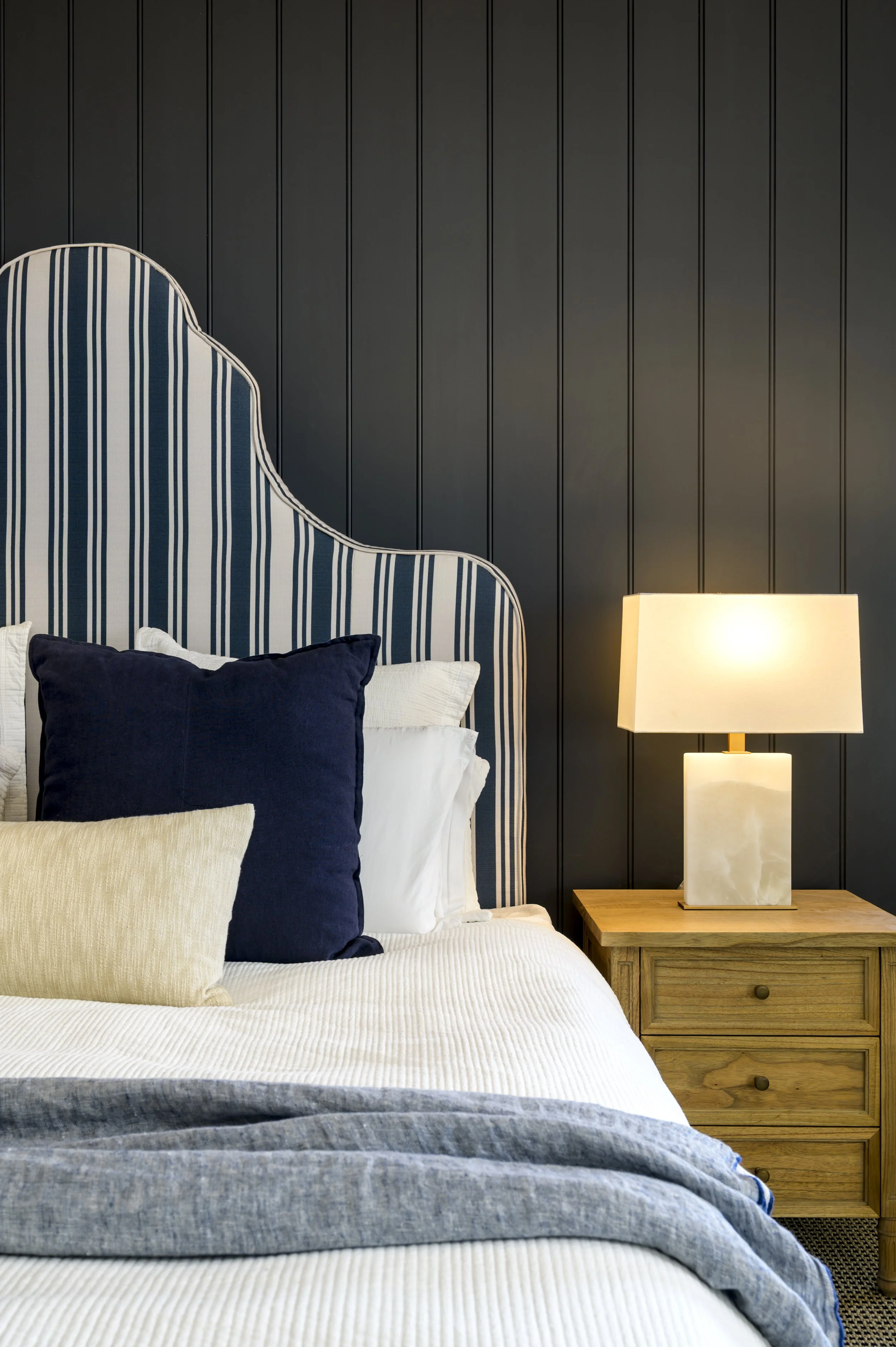 A bedroom with a blue and white striped upholstered bed head, dark textured wall, bedside wooden table, and a cream-colored lamp with a rectangular base.
