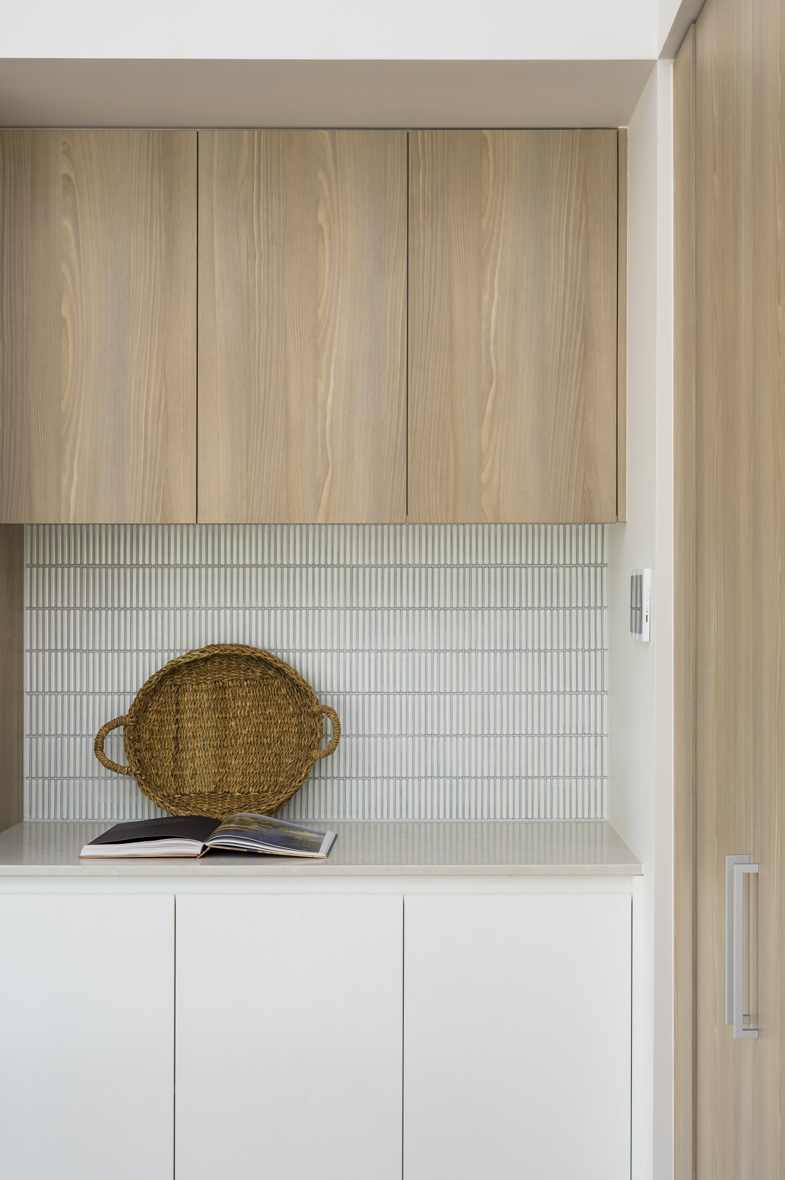 Kitchen cabinet with a woven tray and an open book on the countertop, white tiled backsplash.