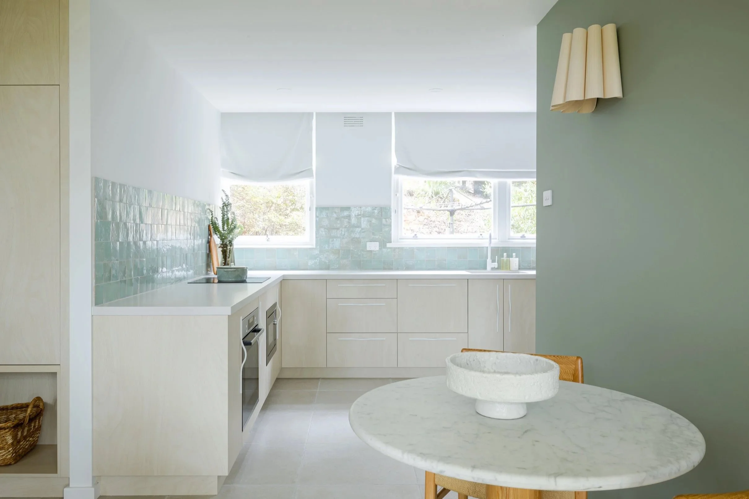 Bright kitchen with white cabinets and countertops, green tiled backsplash, two windows with white blinds, and a marble dining table with a white bowl, decorated in pastel tones.