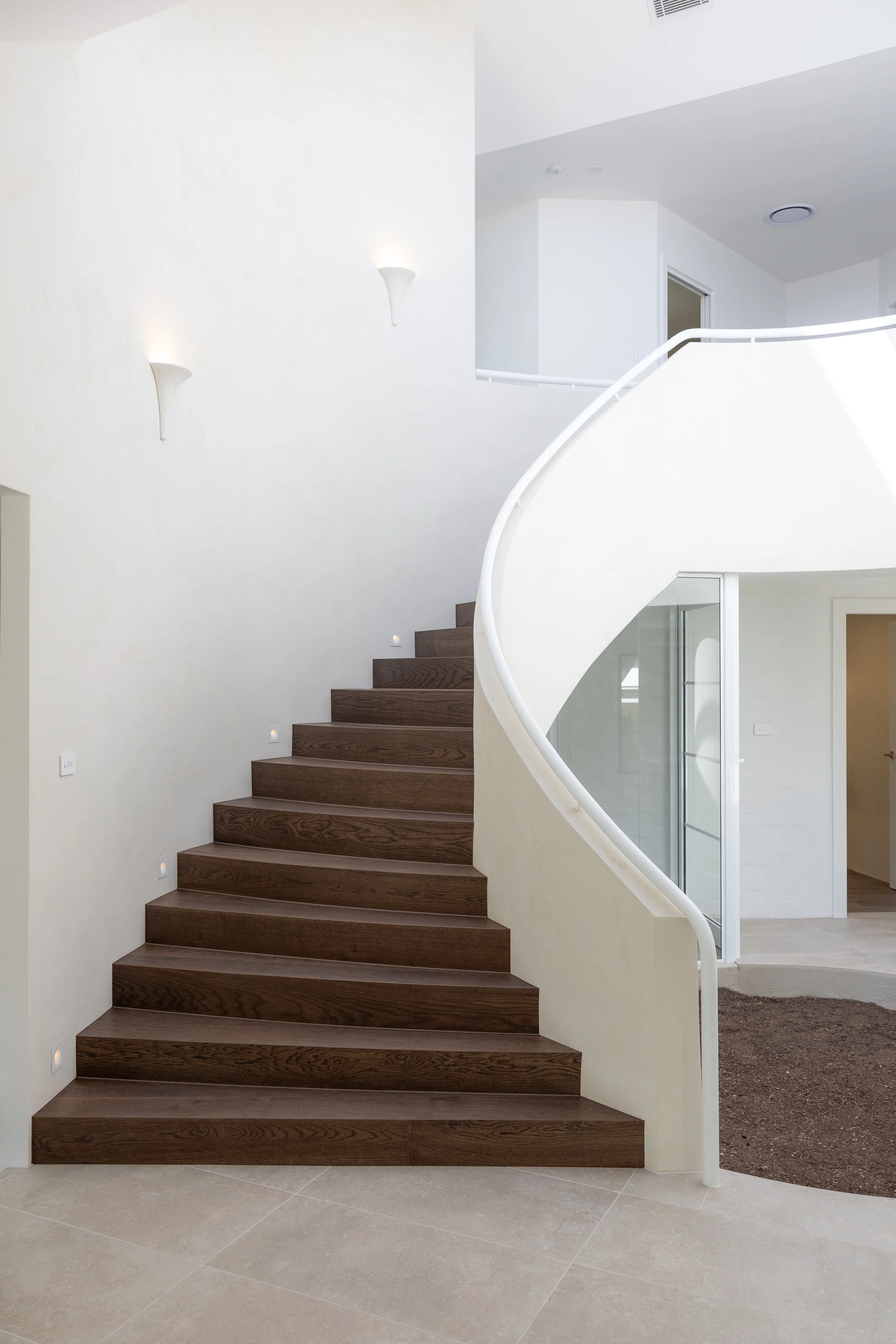 Interior of a modern home featuring a curved staircase with dark wooden steps and a white railing, beige tile flooring, and white walls with wall-mounted lights.
