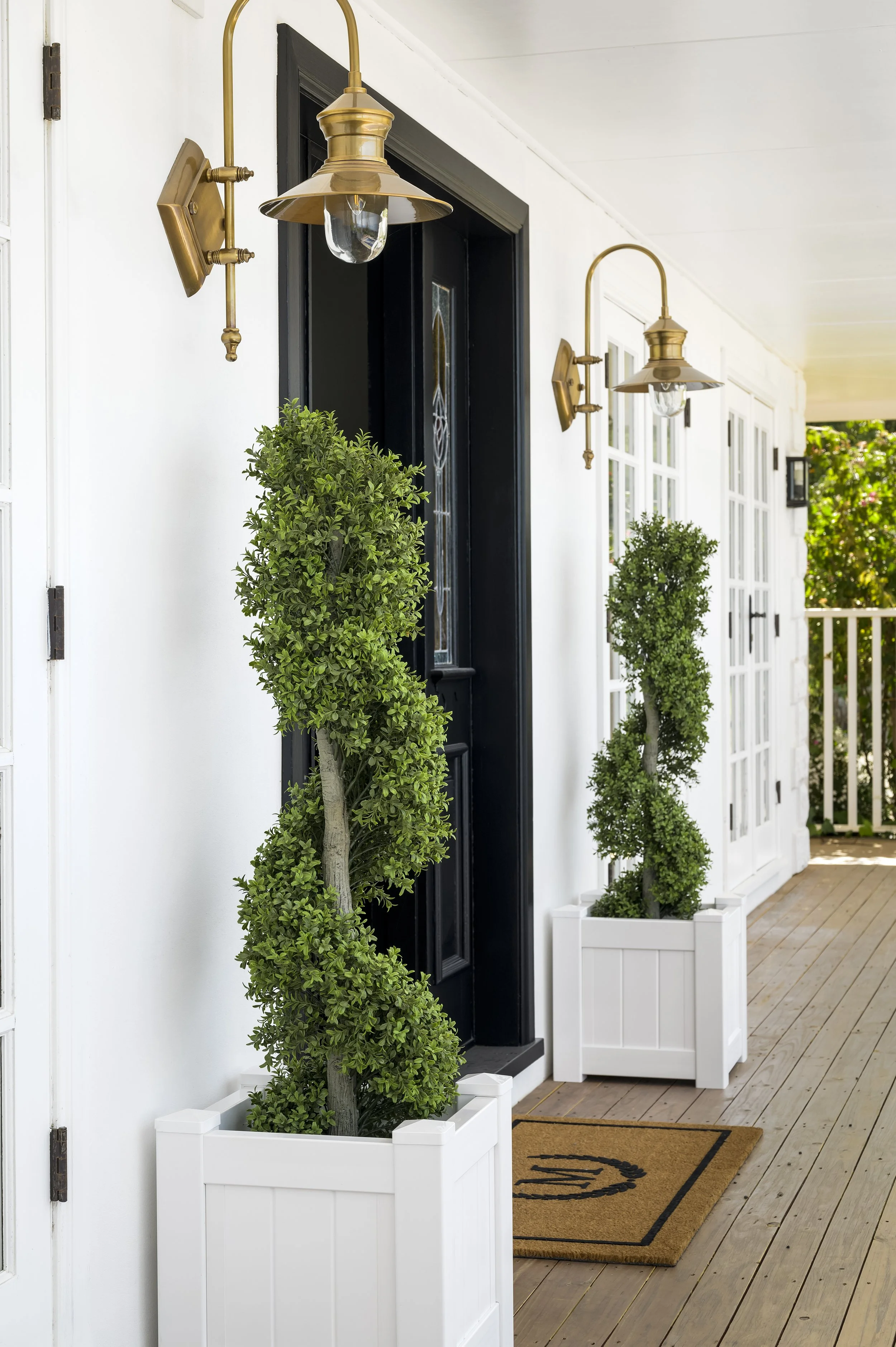 A porch with two black and gold wall-mounted lamps, two twisted topiary plants in white square planters, a welcome mat, and a front door with a glass panel.