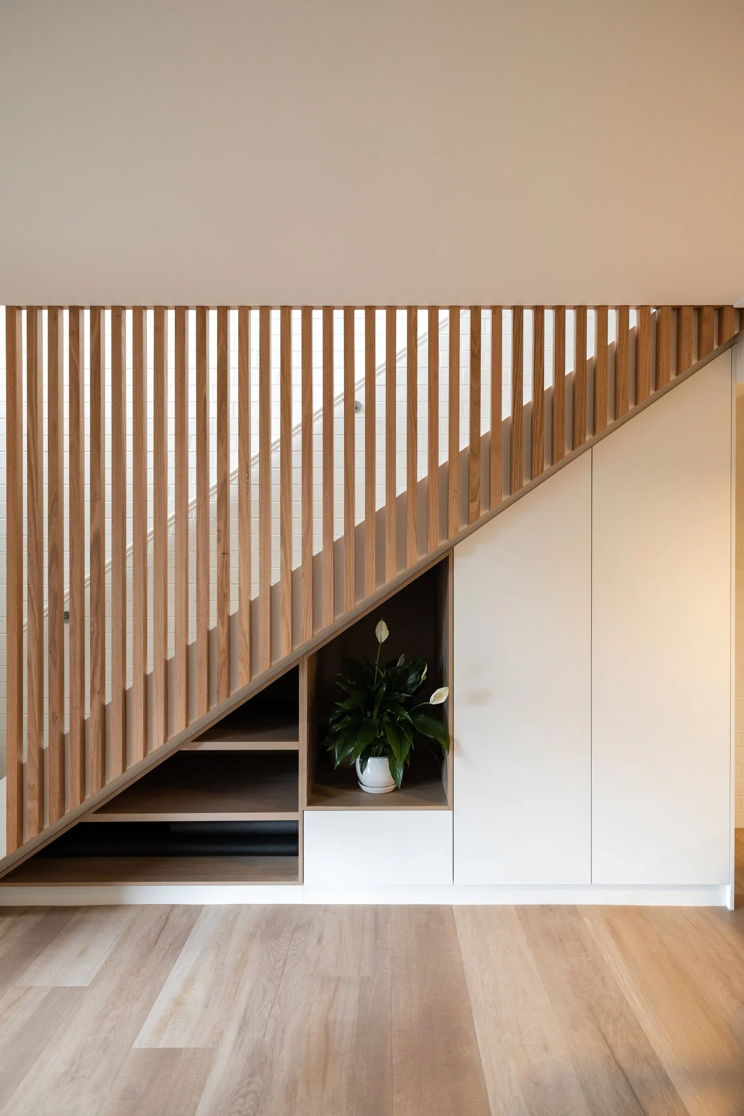 Interior of a modern home showing a staircase with wooden balustrades, built-in storage with a plant, and a light-colored wooden floor.