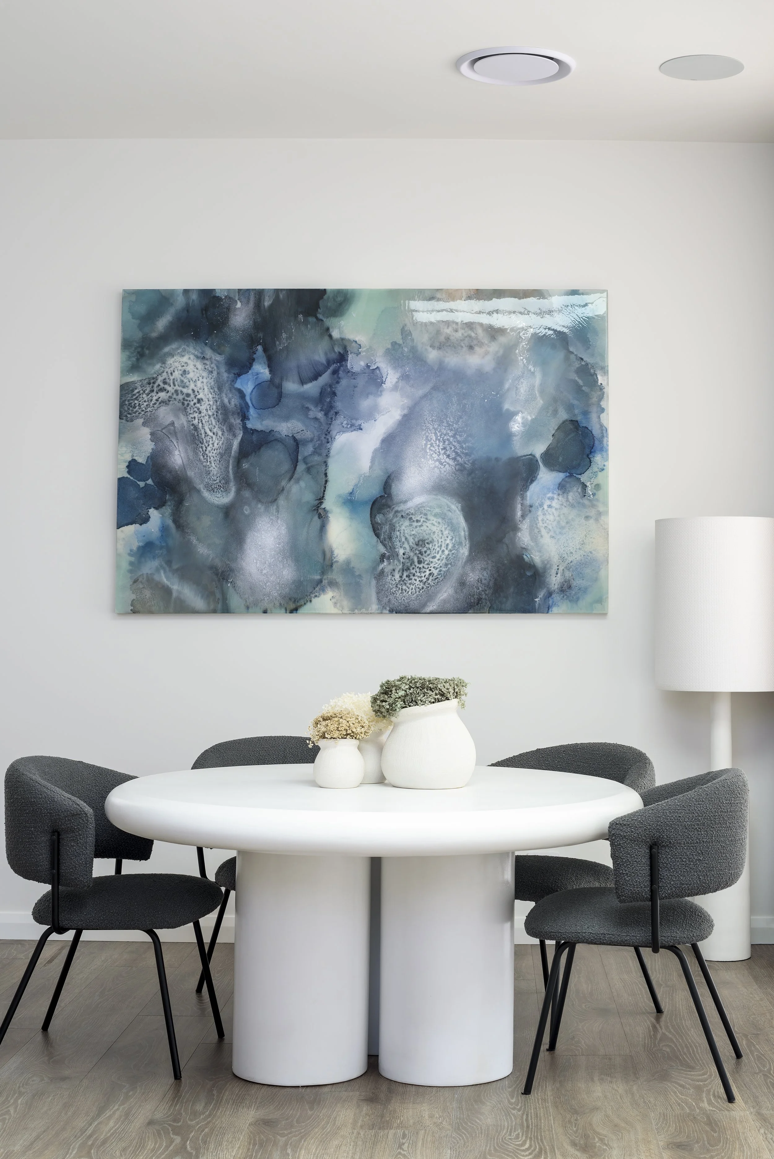Modern dining room with white oval table, four dark gray chairs, large abstract blue and gray wall art, white ceramic vases with flowers, white floor lamp, and light wood flooring.