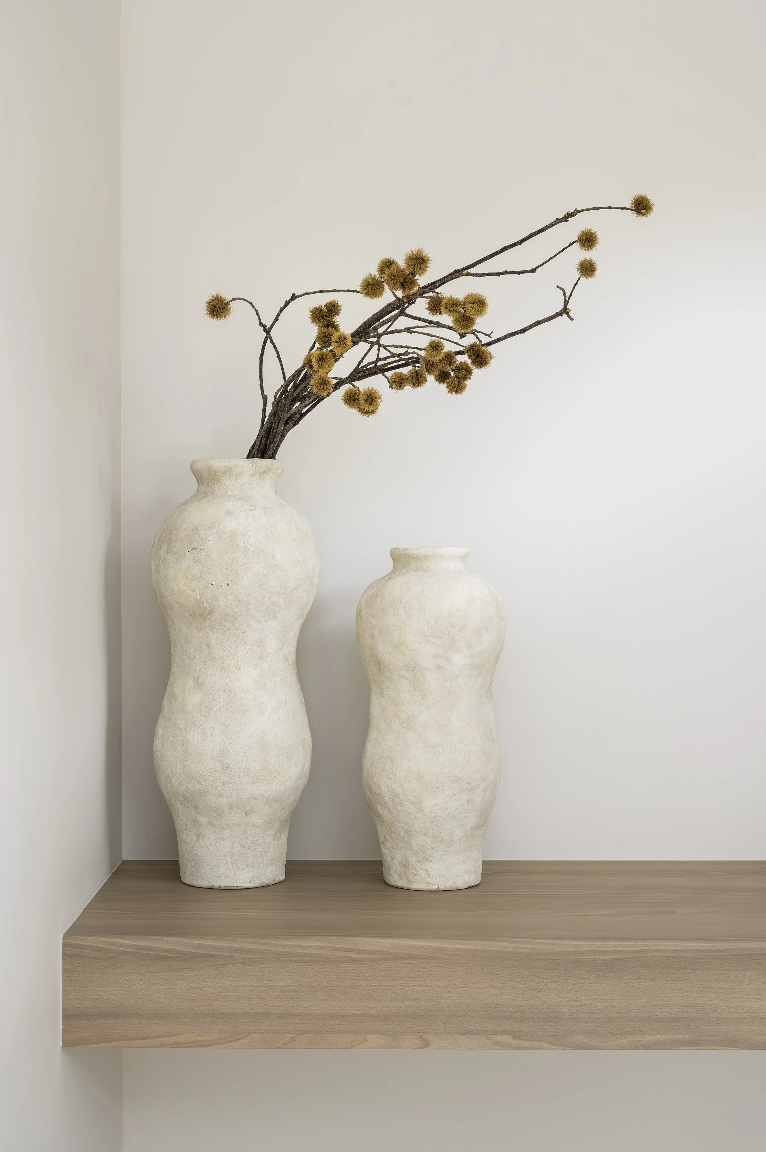 Two beige ceramic vases, one taller than the other, with dried branches and spherical flowers, placed on a wooden shelf against a white wall.
