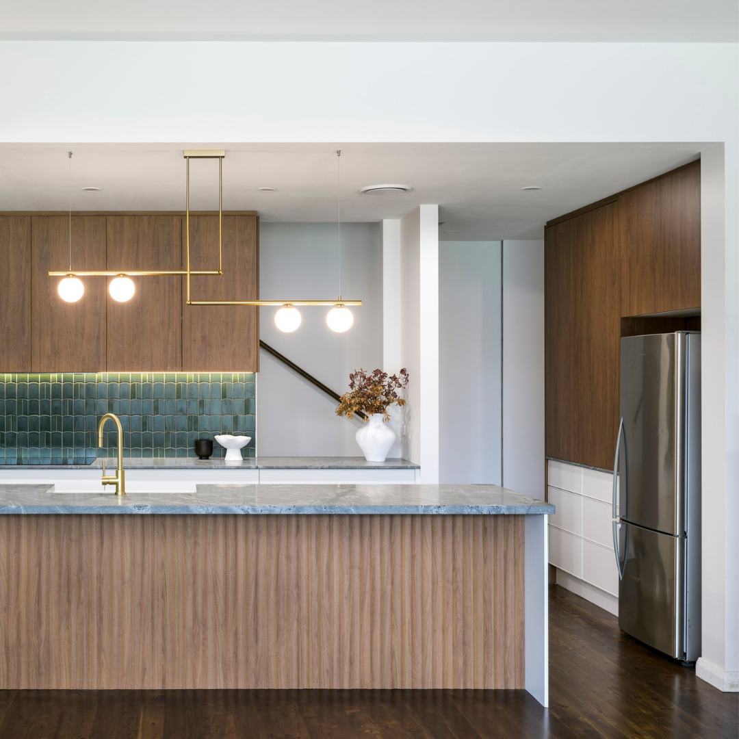 Modern kitchen with wooden cabinets, a marble countertop, a stainless steel refrigerator, and decorative vases on the counter, illuminated by pendant lights.