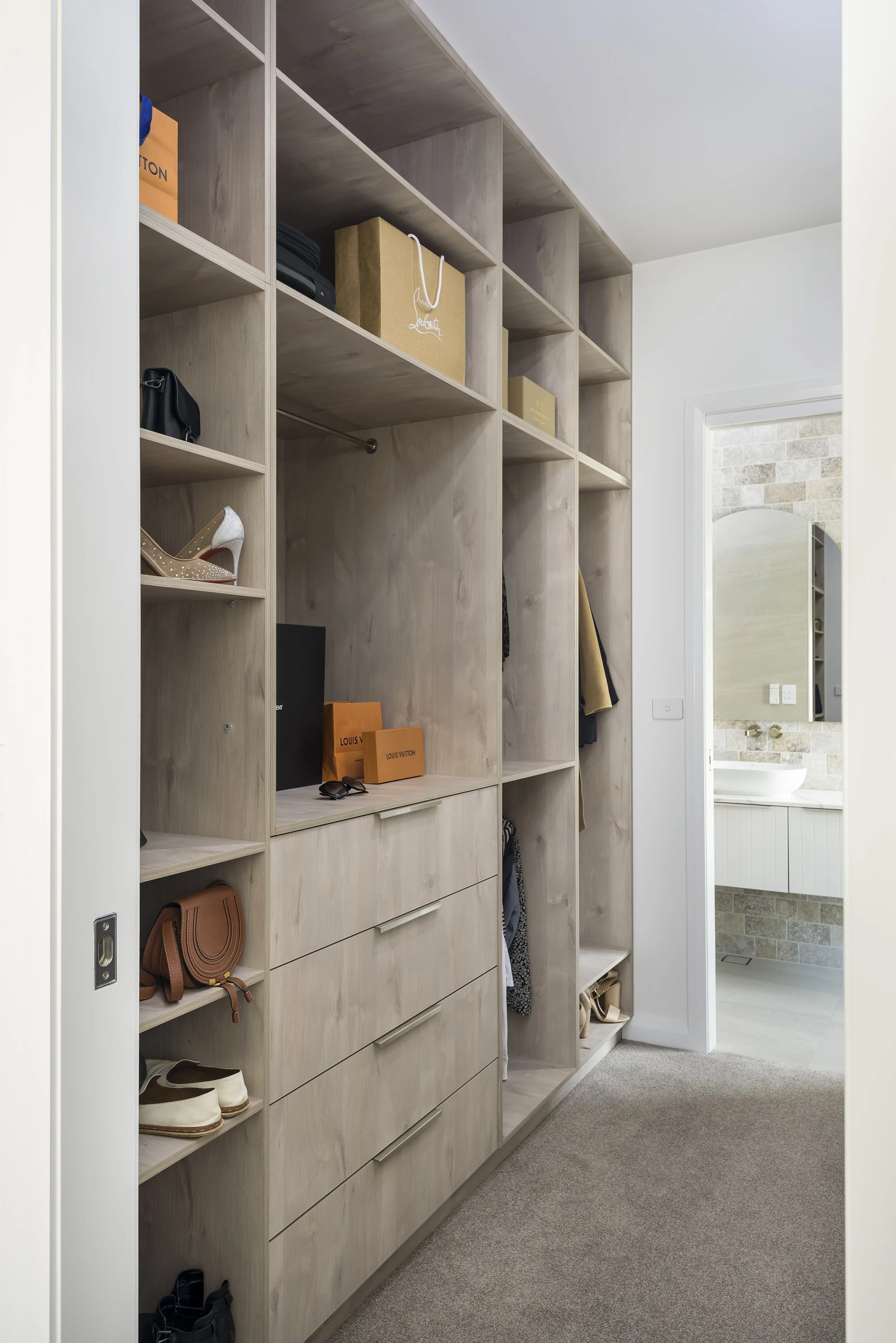 A walk-in closet with light wood shelving and drawers, containing handbags, shoes, and boxes, next to a bathroom with a sink and mirror.
