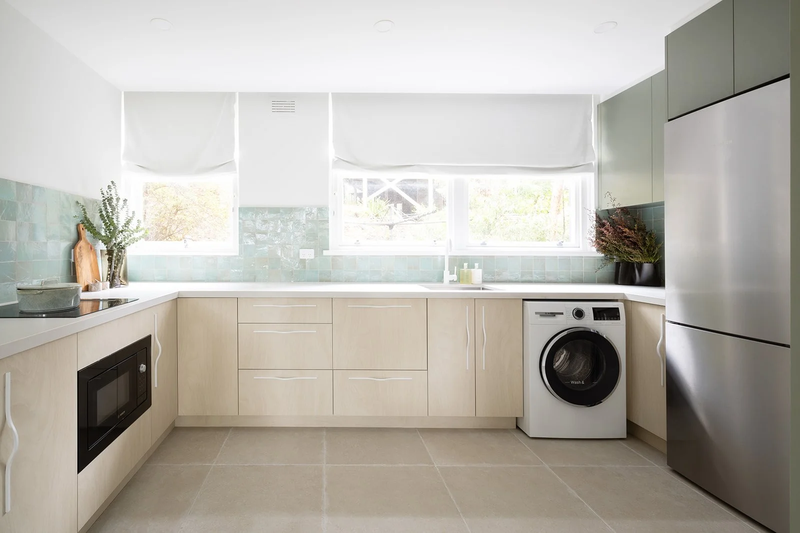 Bright kitchen with light wood cabinets, a refrigerator, microwave, washing machine, and a window with white blinds. Decor includes potted plants and kitchen utensils.