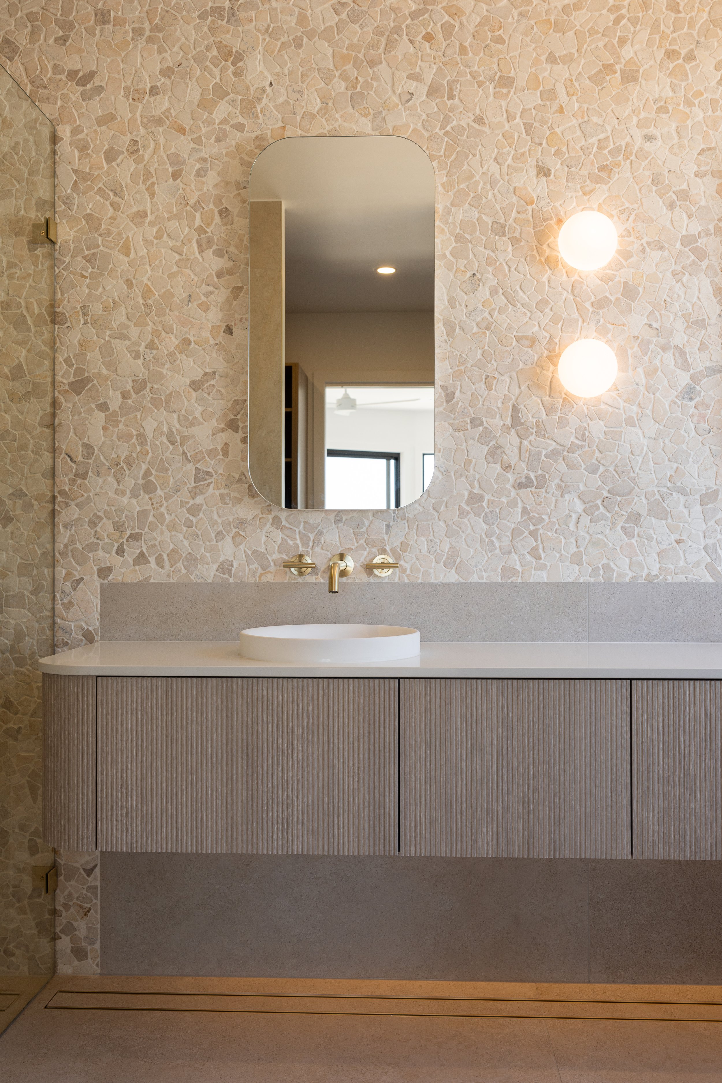 Modern bathroom vanity with a round vessel sink, gold faucet, and a large oval mirror against a textured beige stone wall, illuminated by two round wall sconces.