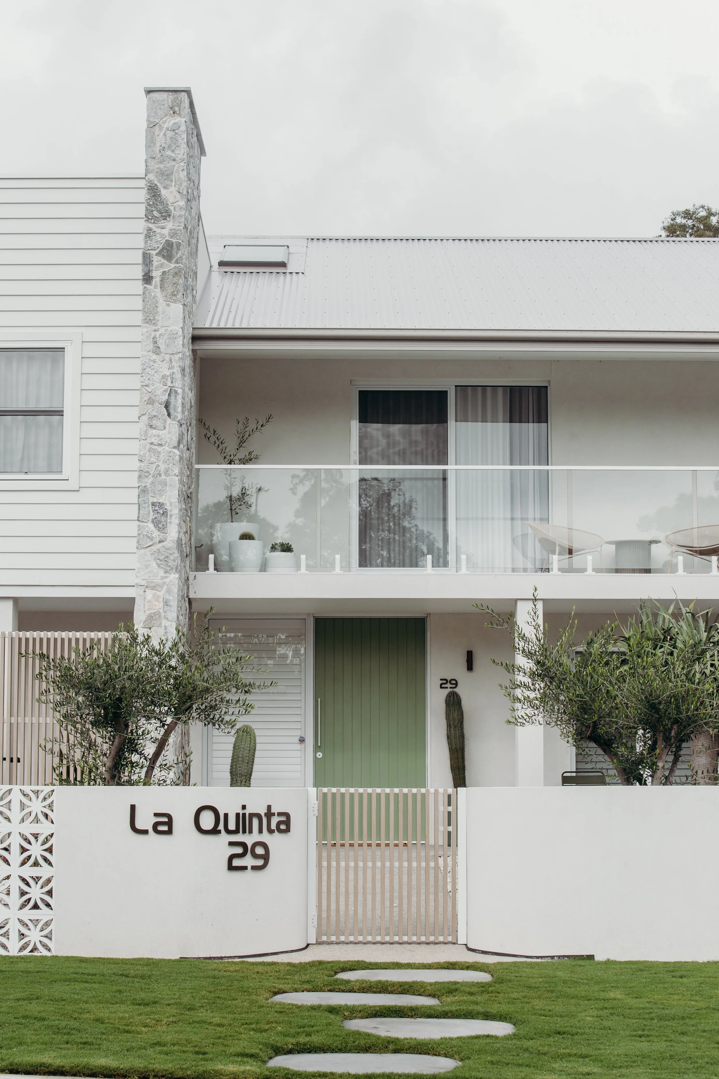 Modern white residential building with a green front door, a white wall fence with the inscription 'La Quinta 29', pathway stones, cacti, and trees in the yard.