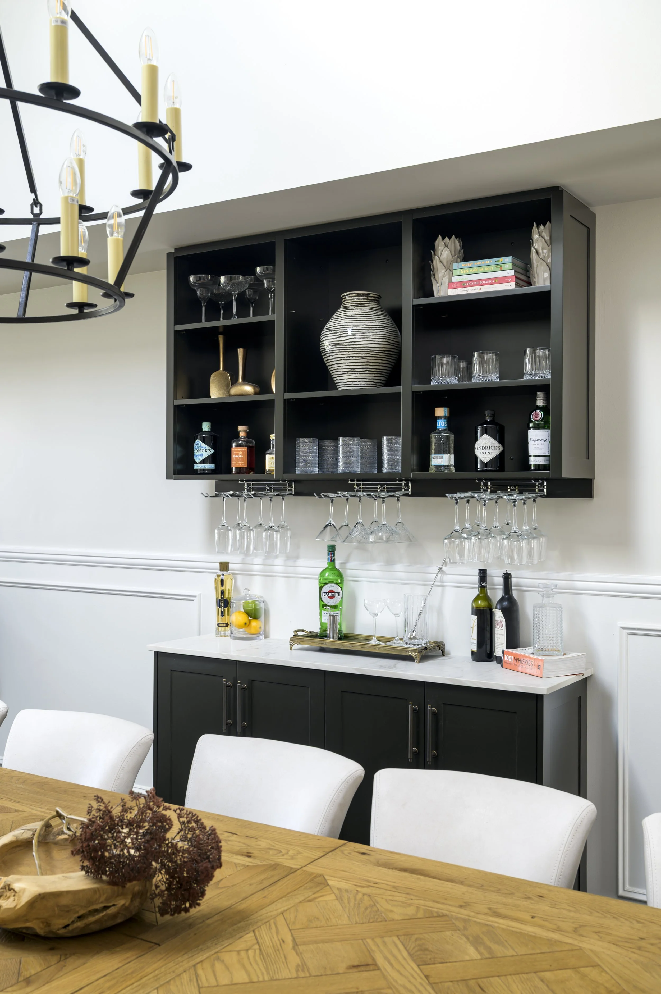 A dining room setup featuring a black cabinet with barware and decorative items, white upholstered chairs around a wooden table, and a black chandelier with candle-like light bulbs.