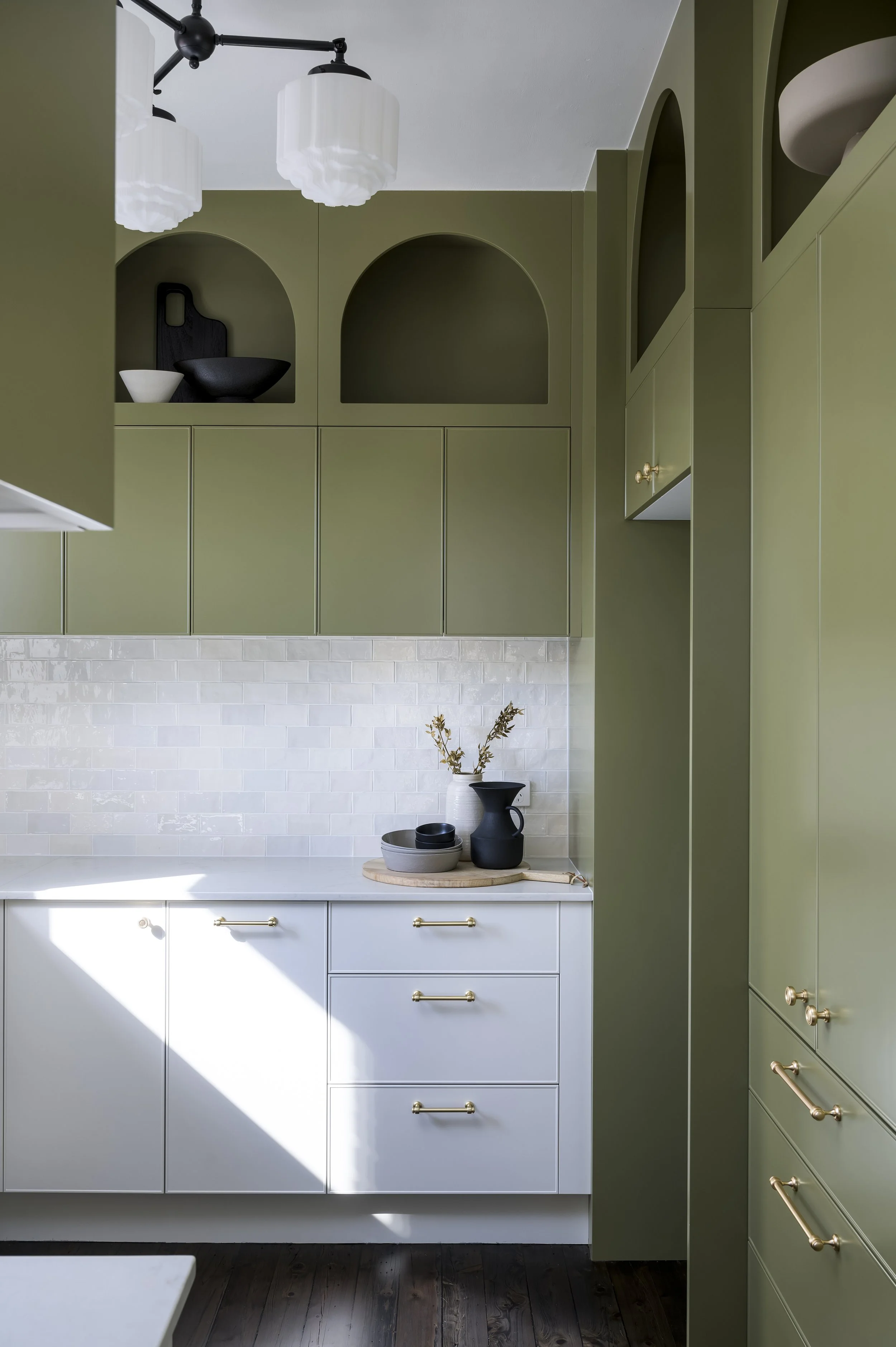 Kitchen with white cabinetry, olive green upper and tall cabinets, white subway tile backsplash, black and white decorative dishes, and a wooden cutting board with black and white vases and dried plant stems.