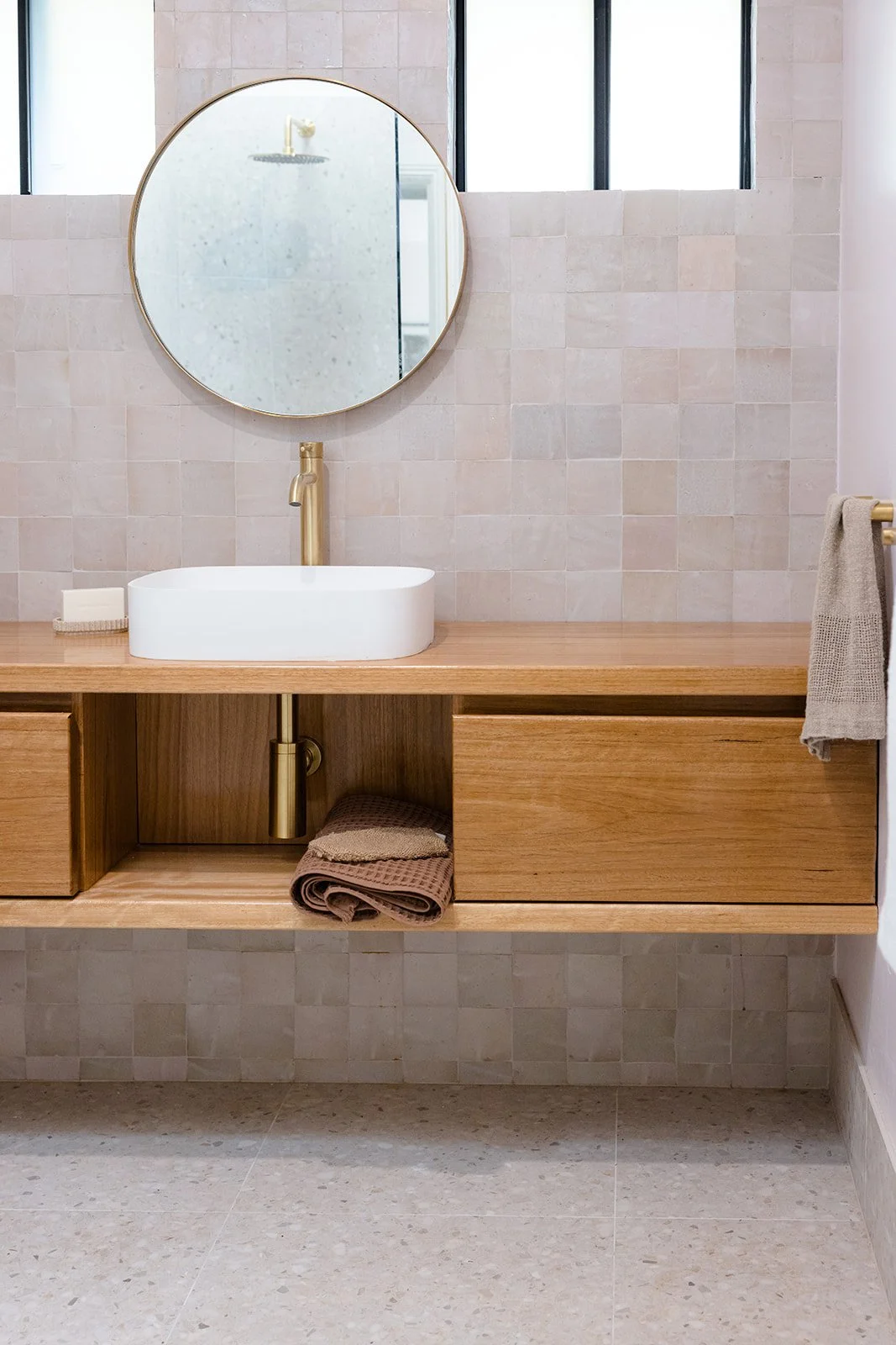 Modern bathroom with a round mirror, white vessel sink, and wooden vanity. Beige tiled wall and floor with a towel hanging on a gold towel rack.