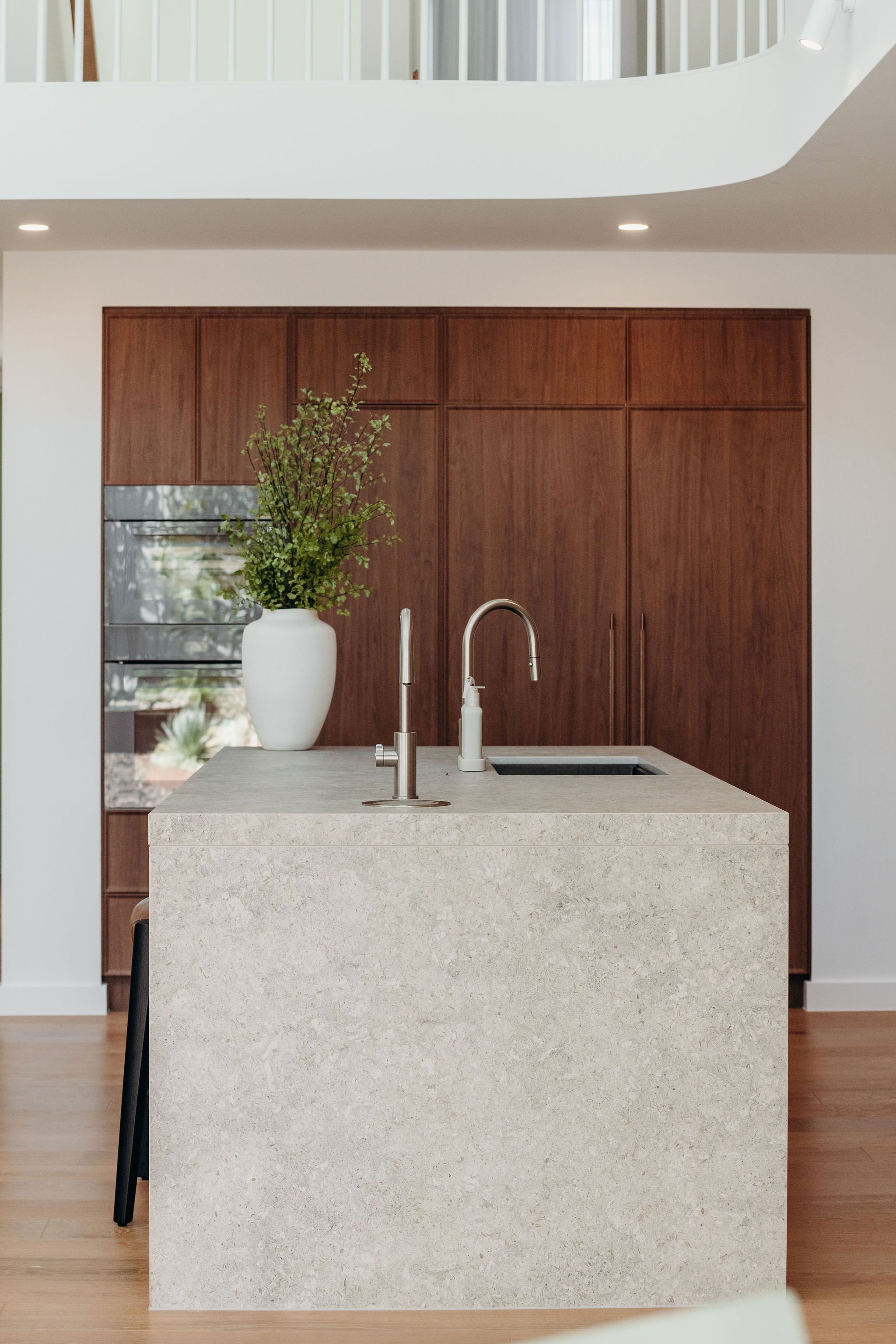 Modern kitchen with a beige stone island, a white vase with greenery, and wooden cabinetry.