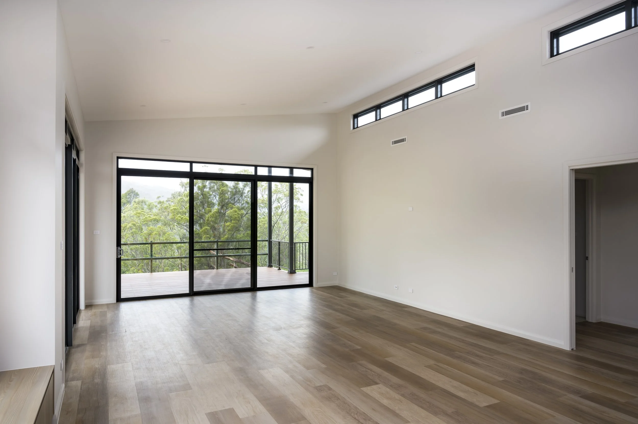 Empty living room with light wood floors, white walls, large sliding glass door leading to a balcony, and multiple small windows high on the wall, overlooking green trees.