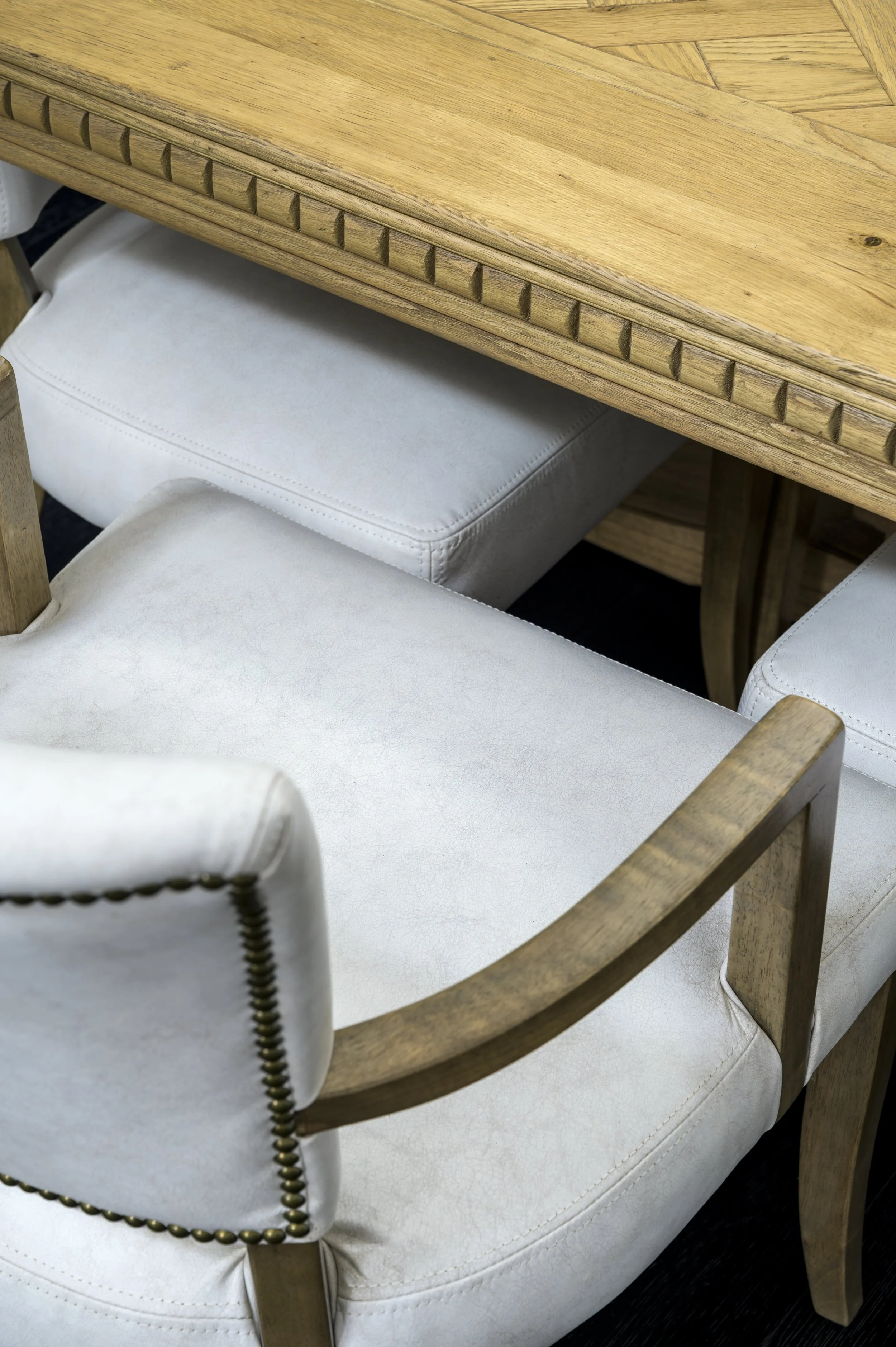 Close-up of a wooden dining table with a natural finish, and upholstered dining chairs with light-colored fabric and nailhead trim.