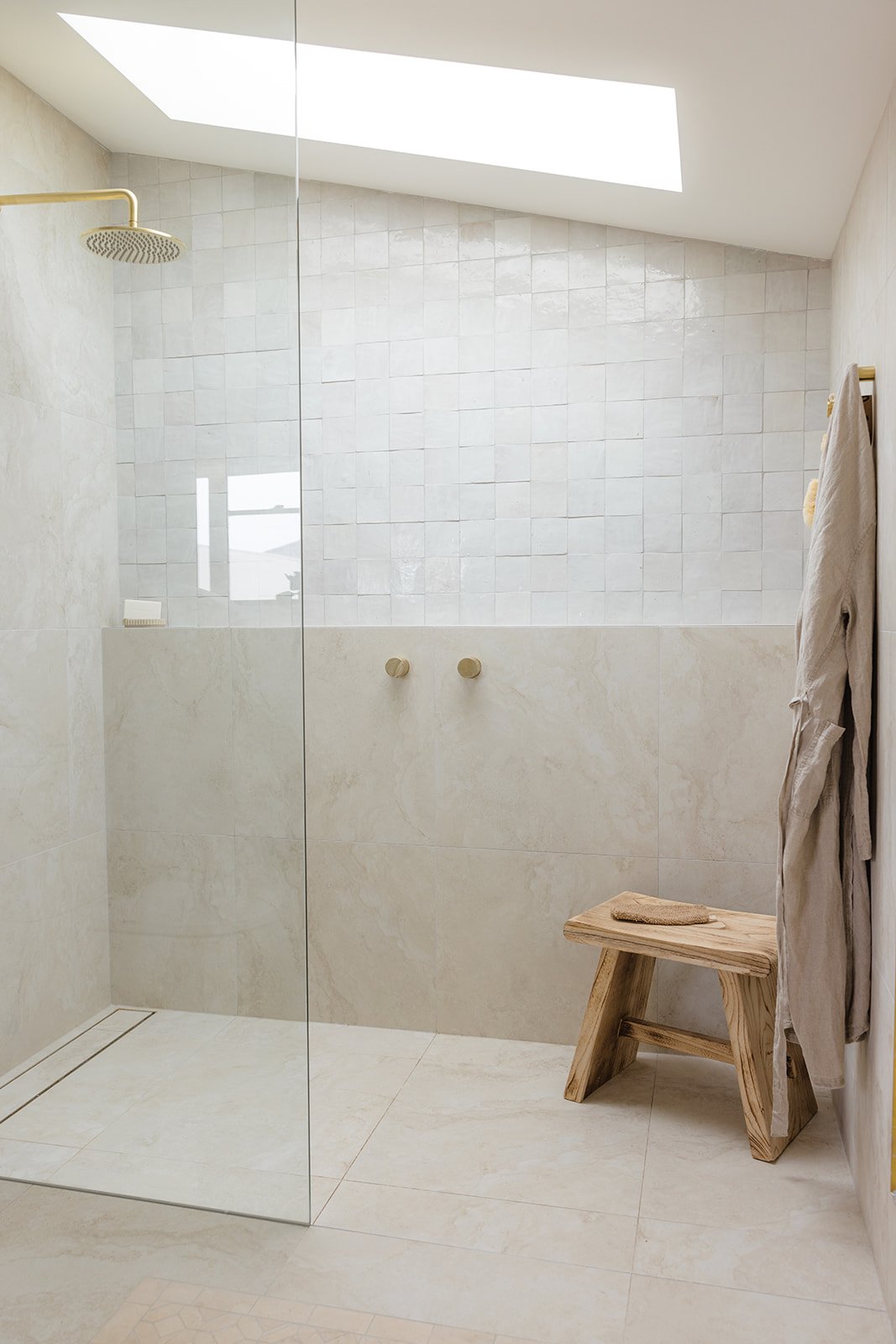 Minimalist bathroom with a walk-in shower, beige tiles, a skylight, a wooden bench, and towels hanging on the wall.