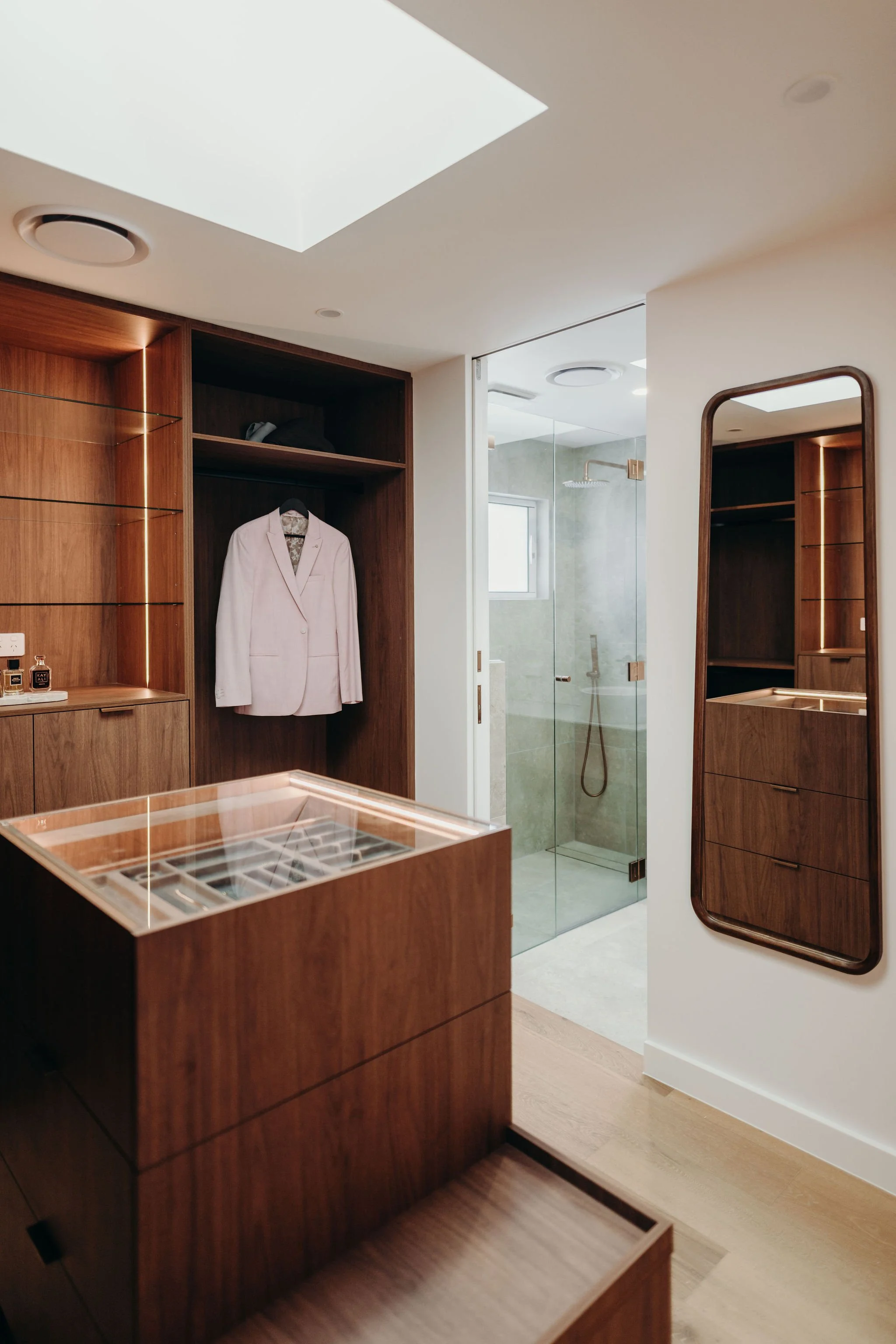 Modern walk-in closet with wooden cabinetry, a mirror, and a glass-enclosed bathroom with a shower visible in the background.