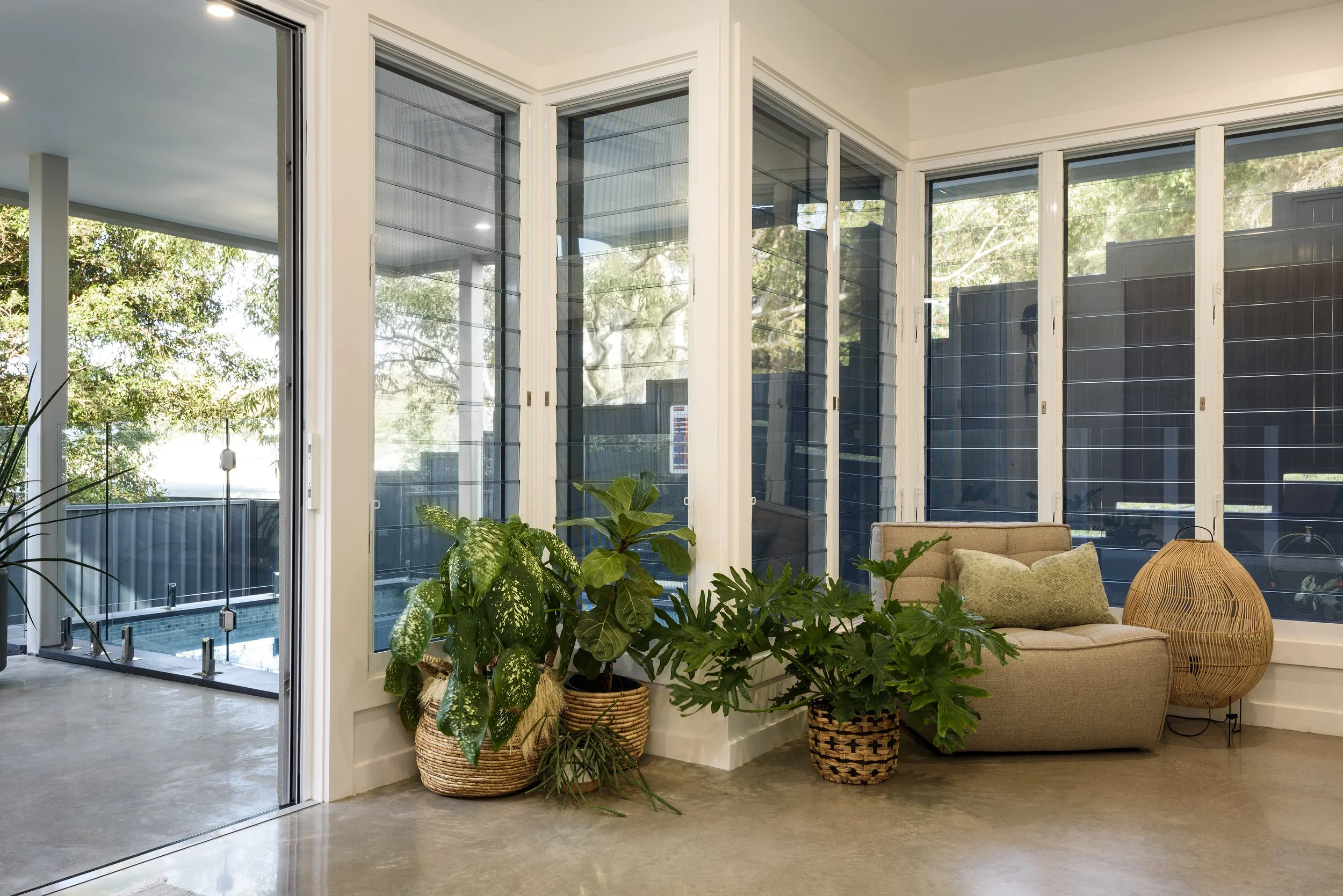 Sunroom with large windows, potted plants, and a beige upholstered chair with pillows, next to a rattan lamp.