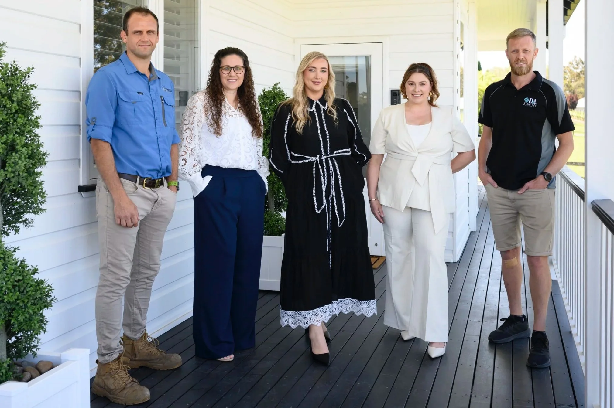 Five people standing on a porch, posing for a photo. The group includes three women and two men, all dressed in various outfits, with a white house and green bushes in the background.