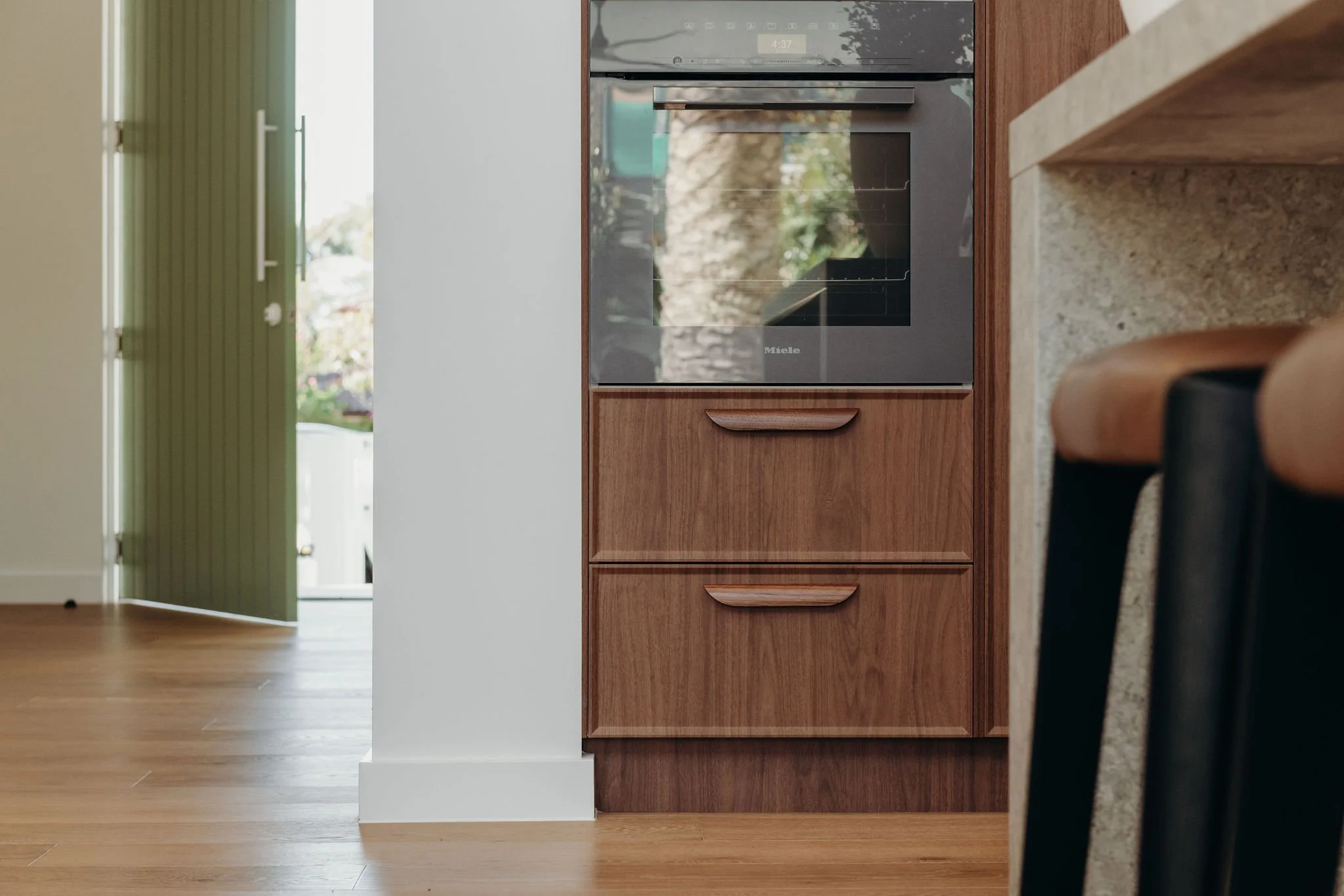 Close-up of a built-in oven in a kitchen with wooden cabinetry, countertop, and a black appliance below the oven. The background shows an open door leading outside with greenery visible.