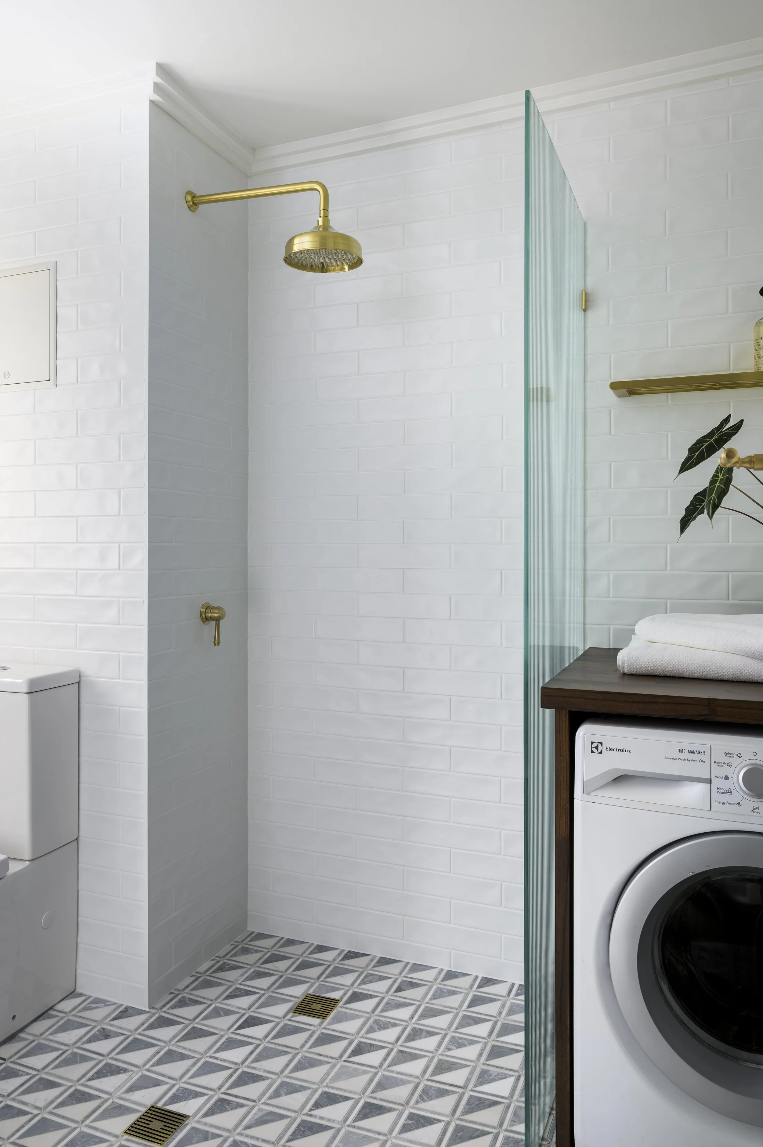 Modern bathroom with a white brick wall, gold shower head, clear glass partitions, and a wooden cabinet with a washing machine.