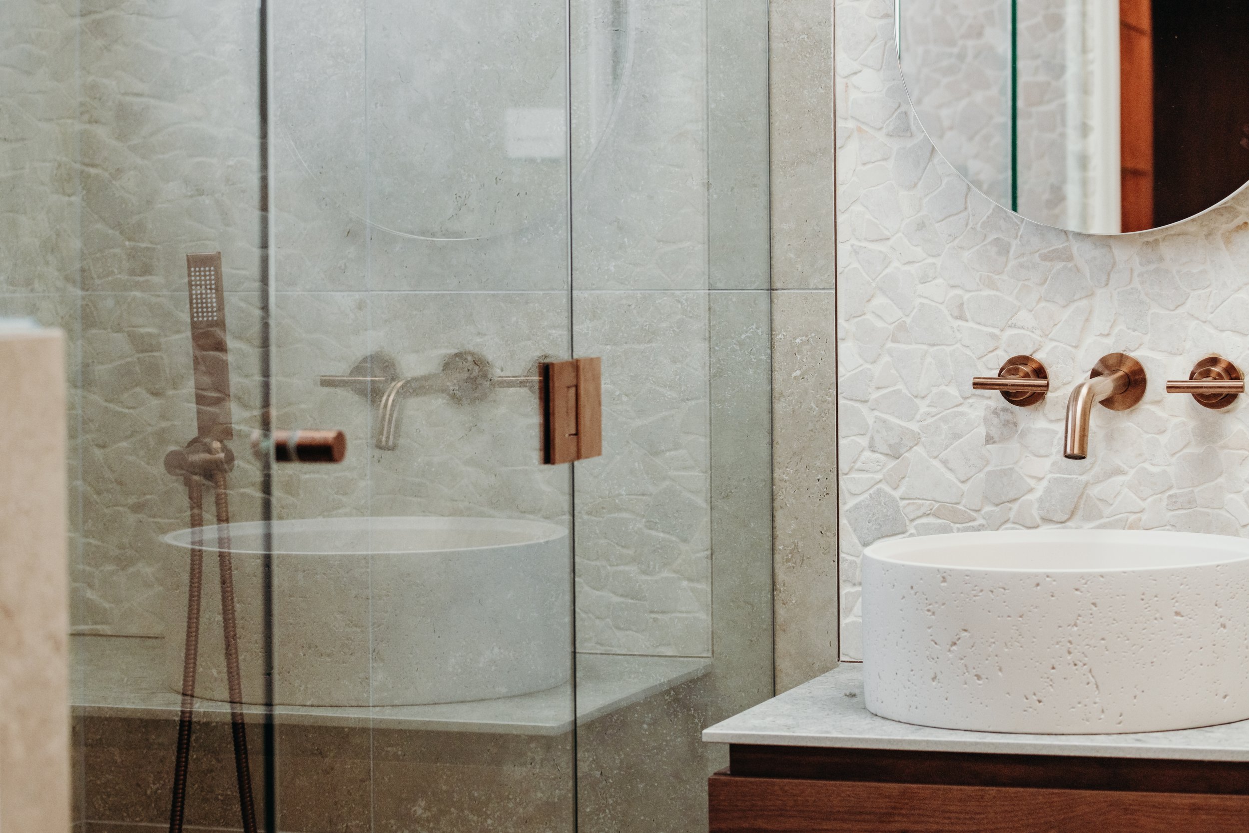 Modern bathroom with a round vessel sink on a wooden vanity, copper fixtures, and a mirror with textured wall behind