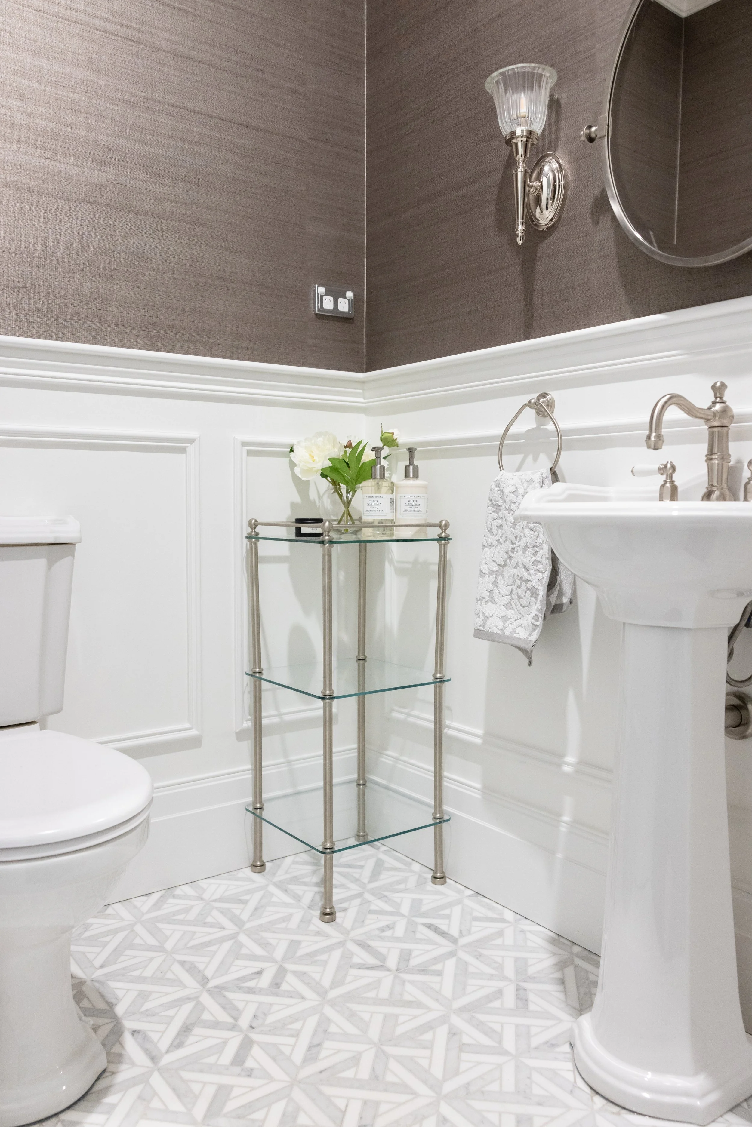 A bathroom with a white toilet, pedestal sink, and a three-tier glass and metal shelf holding toiletries and a flower vase. The wall has a textured dark brown upper half and white lower half with decorative molding, a wall sconce, and a mirror.
