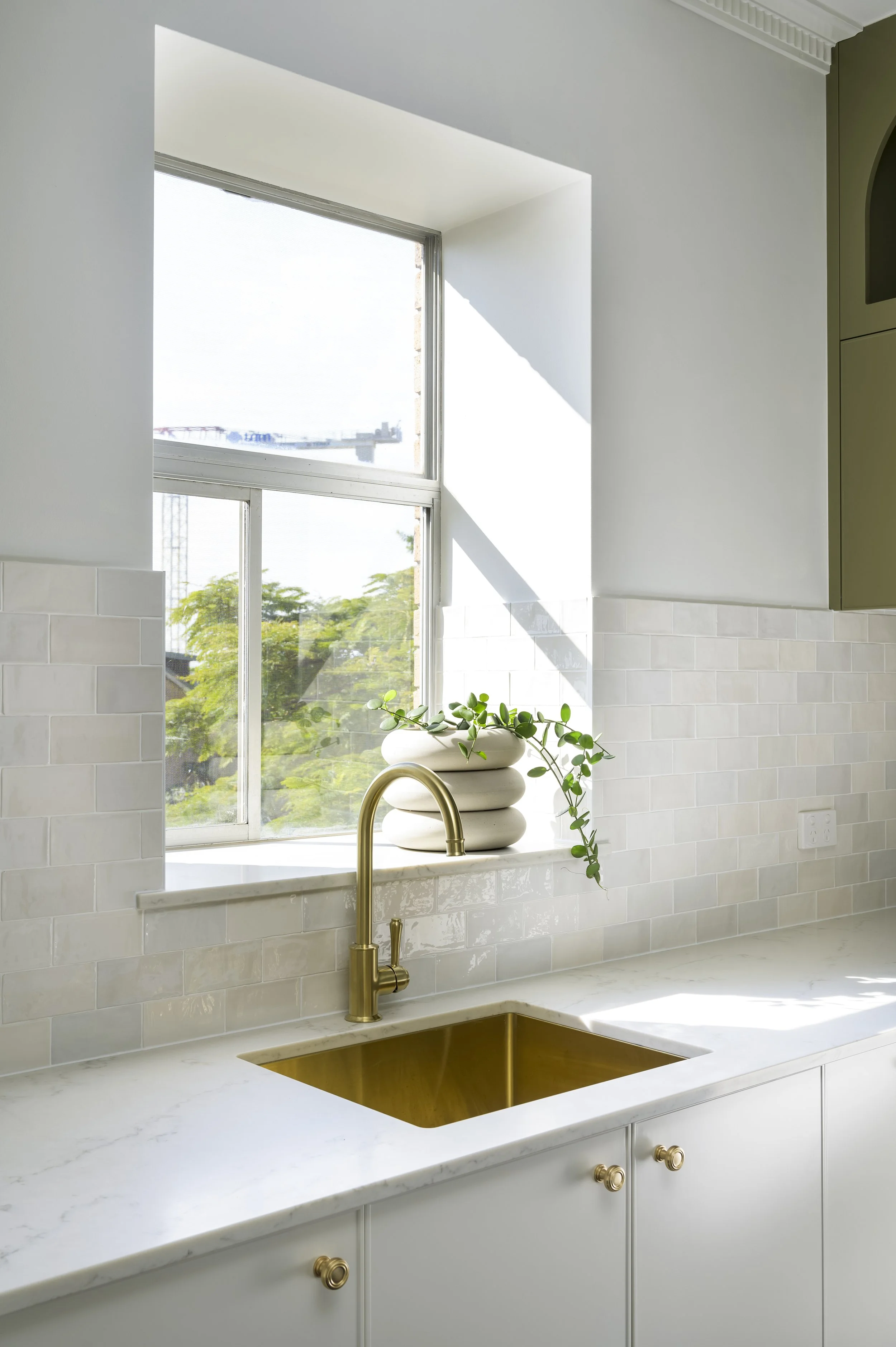Modern kitchen with white cabinetry, marble countertop, gold sink and faucet, and a window with a potted plant on the sill.