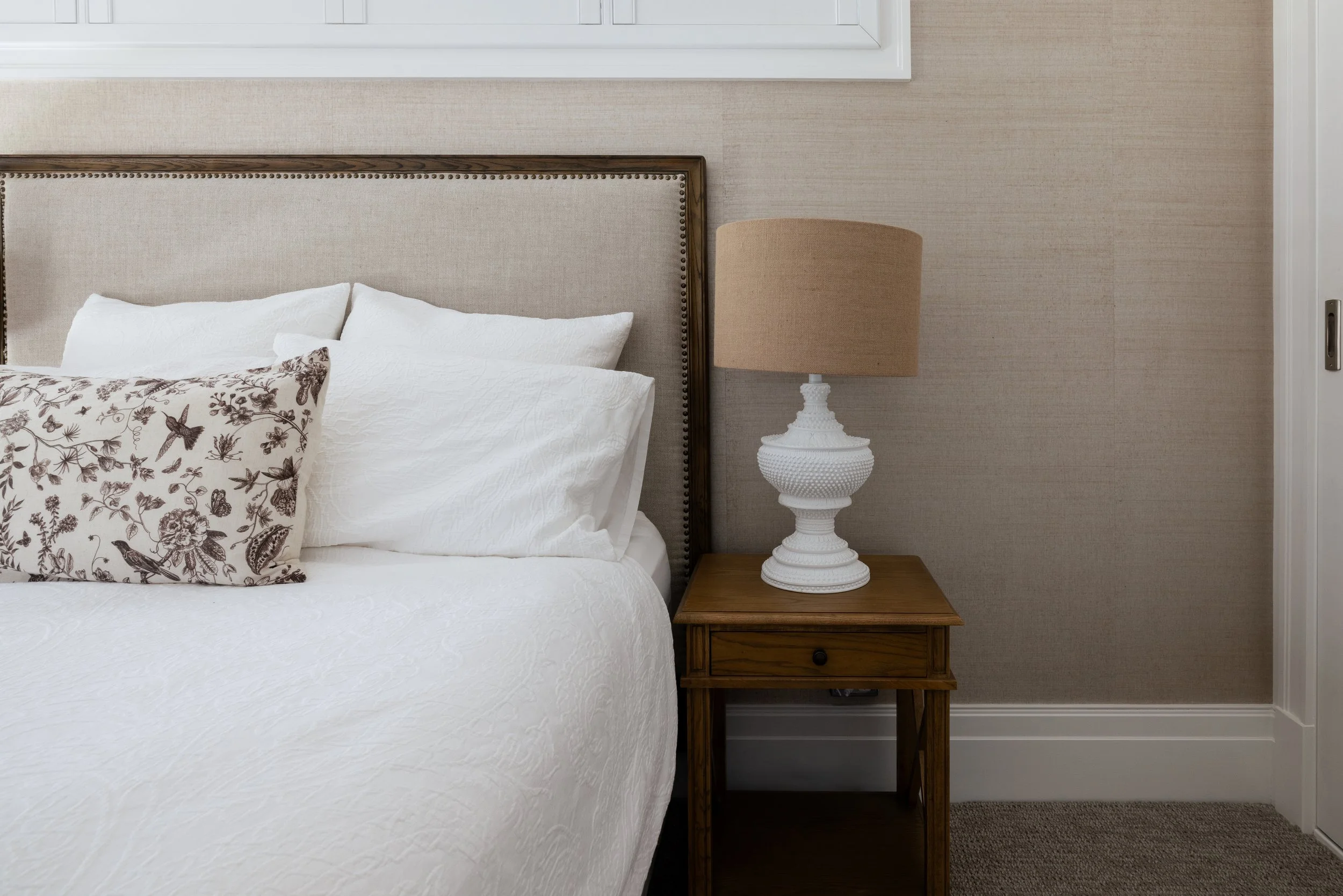 A bedroom with a bed, white bedding, and a decorative pillow with bird and floral design. A wooden nightstand with a white textured lamp and beige lampshade is beside the bed, with a beige wall and closed door in the background.