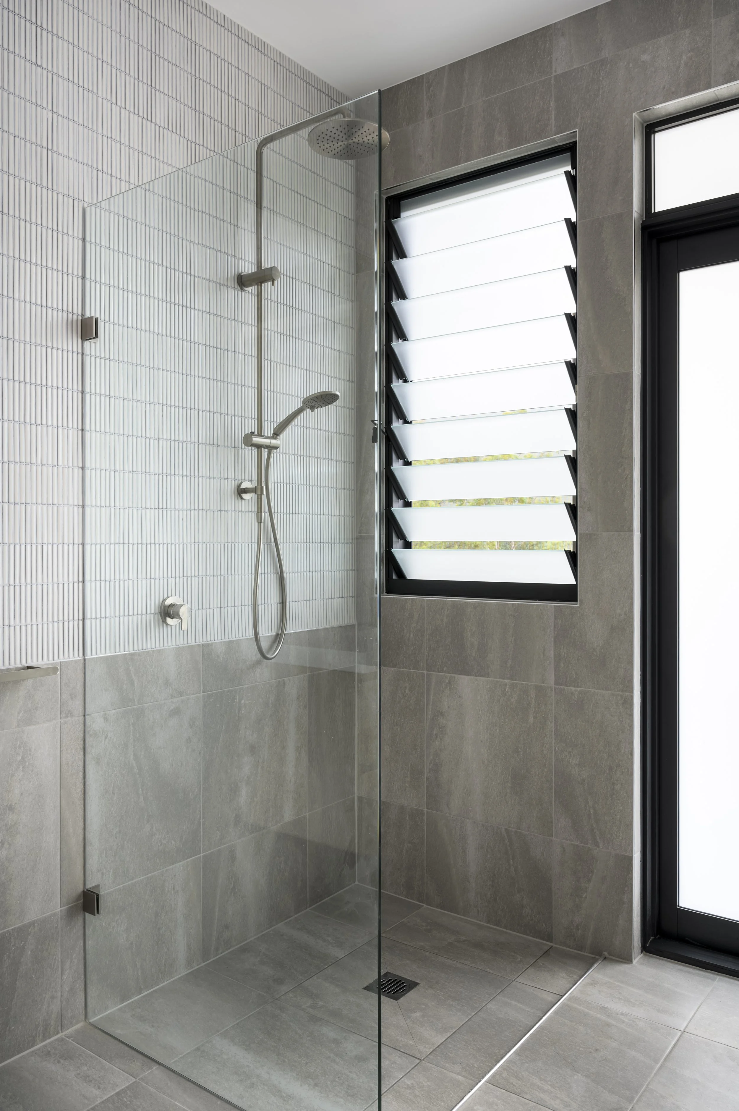 Modern shower with glass wall, handheld showerhead, and large window with adjustable louvered shutters in a tiled bathroom.