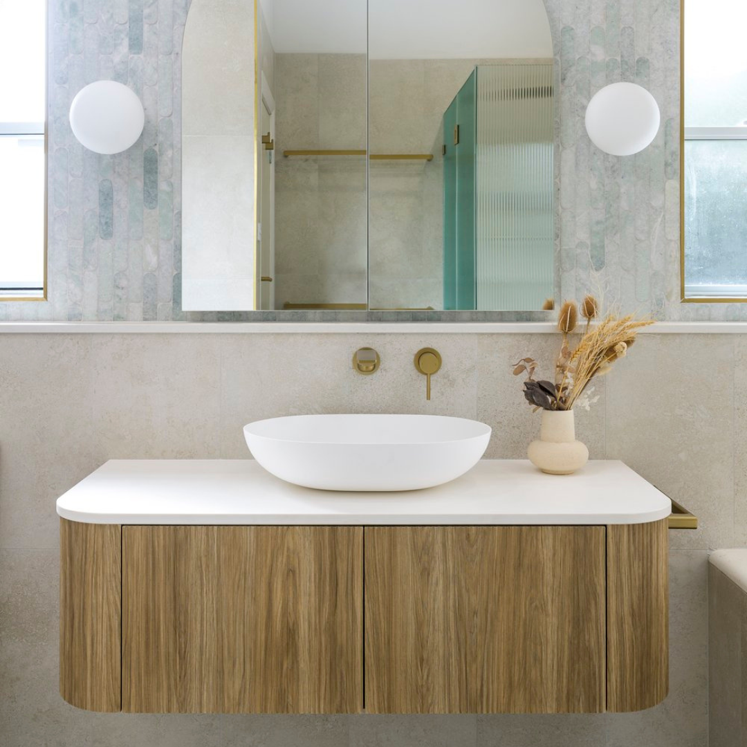 Modern bathroom with a white vessel sink on a wooden vanity, a large mirror, and dried plant arrangement in a vase. Two windows provide natural light.