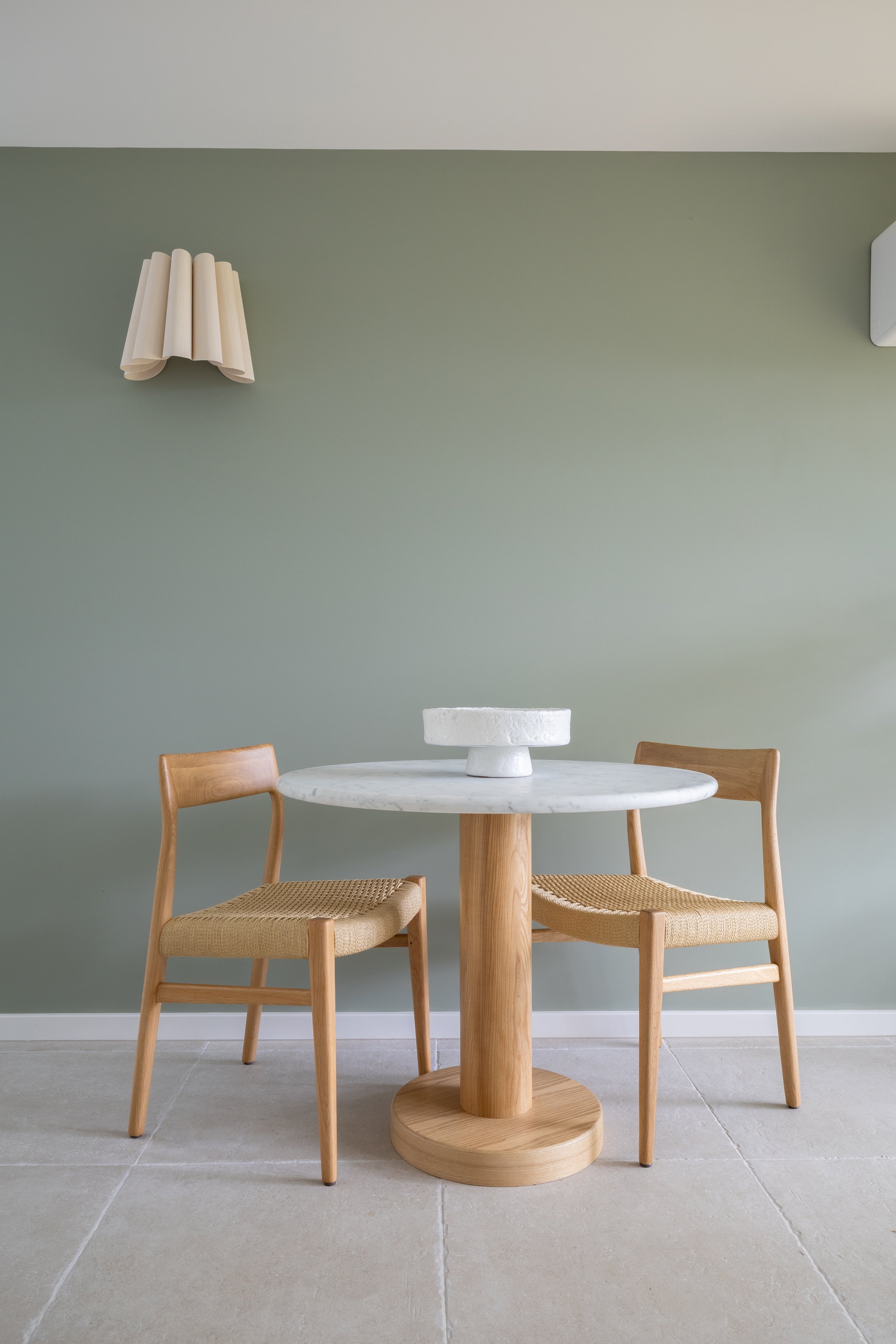 A minimalist dining area with a round white marble table, two wooden chairs with woven seats, a light green wall, a modern wall-mounted light fixture, and a small white vent on the upper right corner.