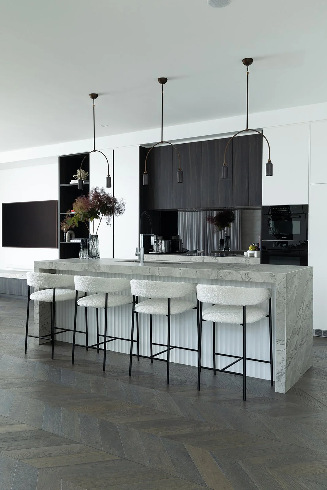 Modern kitchen with white marble island, four white chairs with black legs, black hanging pendant lights, black cabinetry, and a wall-mounted TV.