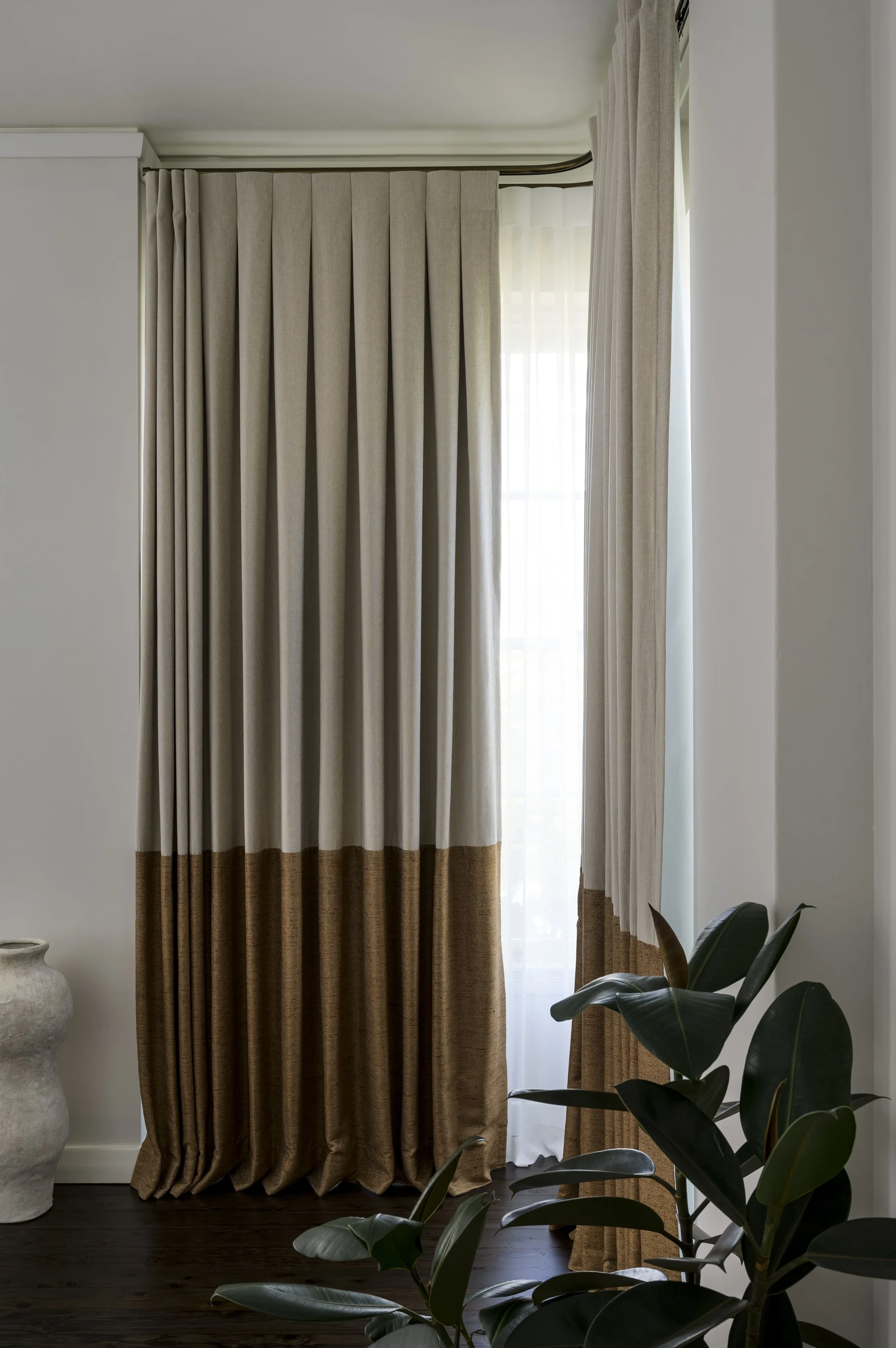 A window with beige and brown curtains, sheer white curtains behind, and a green plant in the foreground.