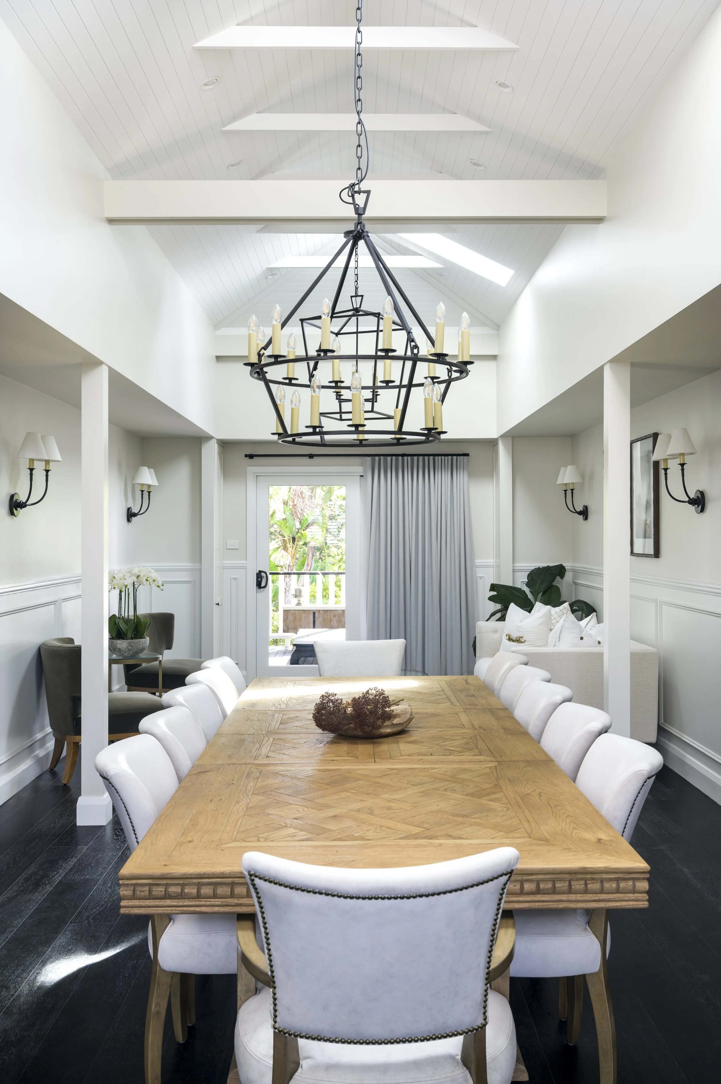 Bright dining room with a large wooden table, white upholstered chairs, a black chandelier, and wall-mounted sconces. A door leads to a balcony with potted plants, and light filters through skylights and a curtain.