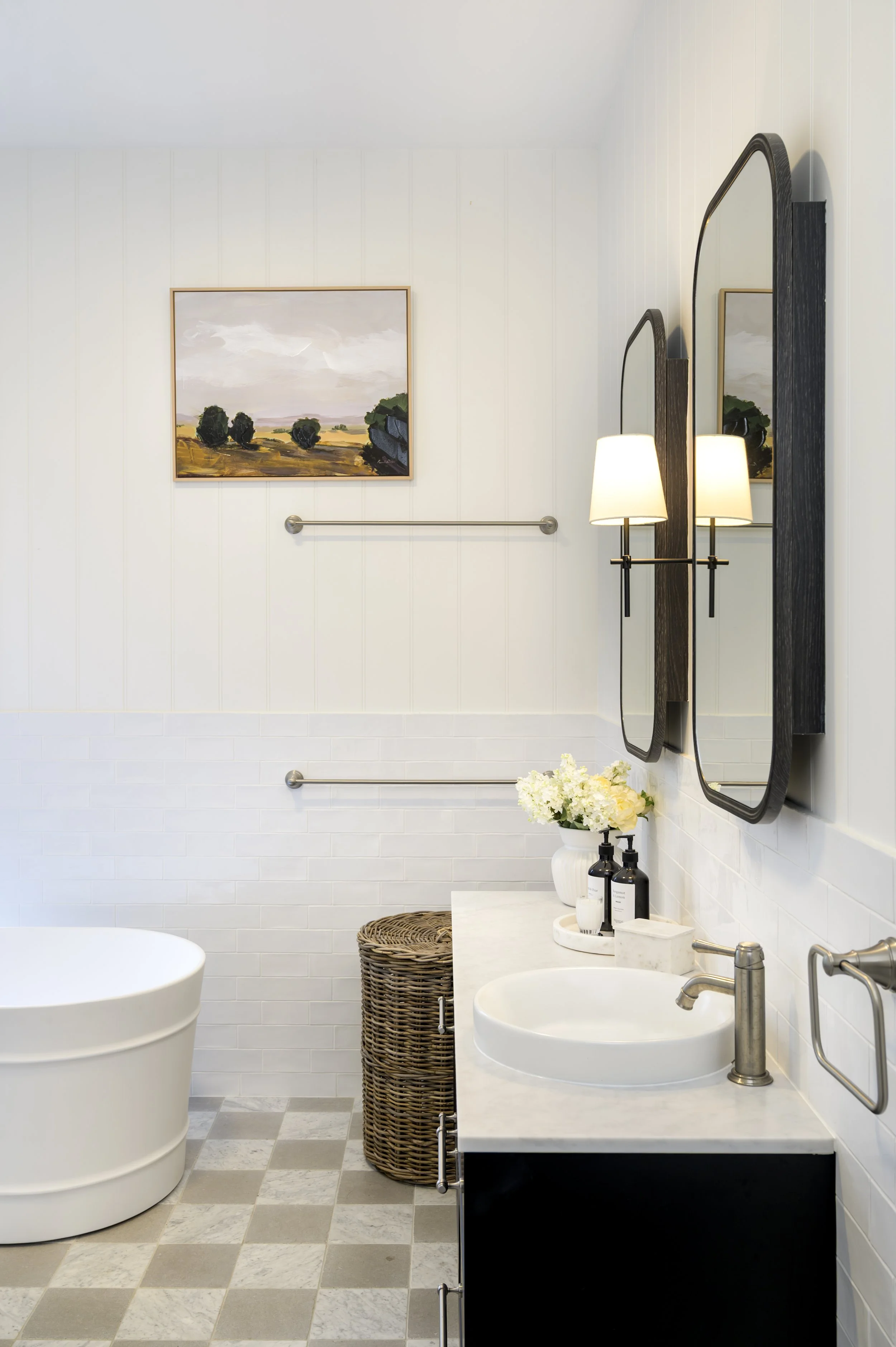 Modern bathroom with white tiled walls, a large white bathtub, a black vanity with a white countertop, a round vessel sink, two mirrors with mounted lights, a flower vase, and a wicker laundry basket.
