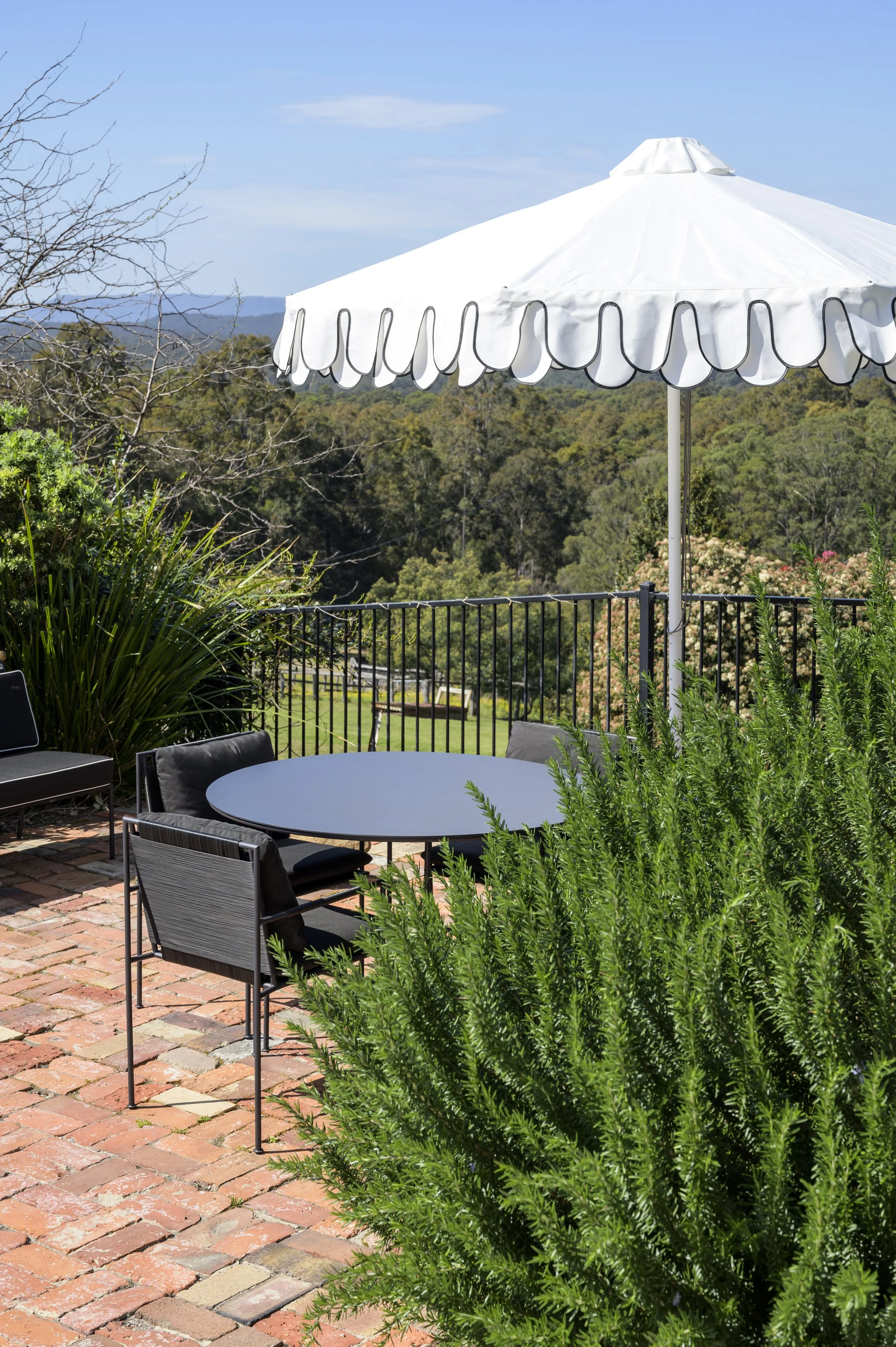 An outdoor patio area with a round black table, black chairs, and a large white patio umbrella. The patio has a brick surface and is surrounded by lush greenery, overlooking a landscape of trees and distant hills under a blue sky.