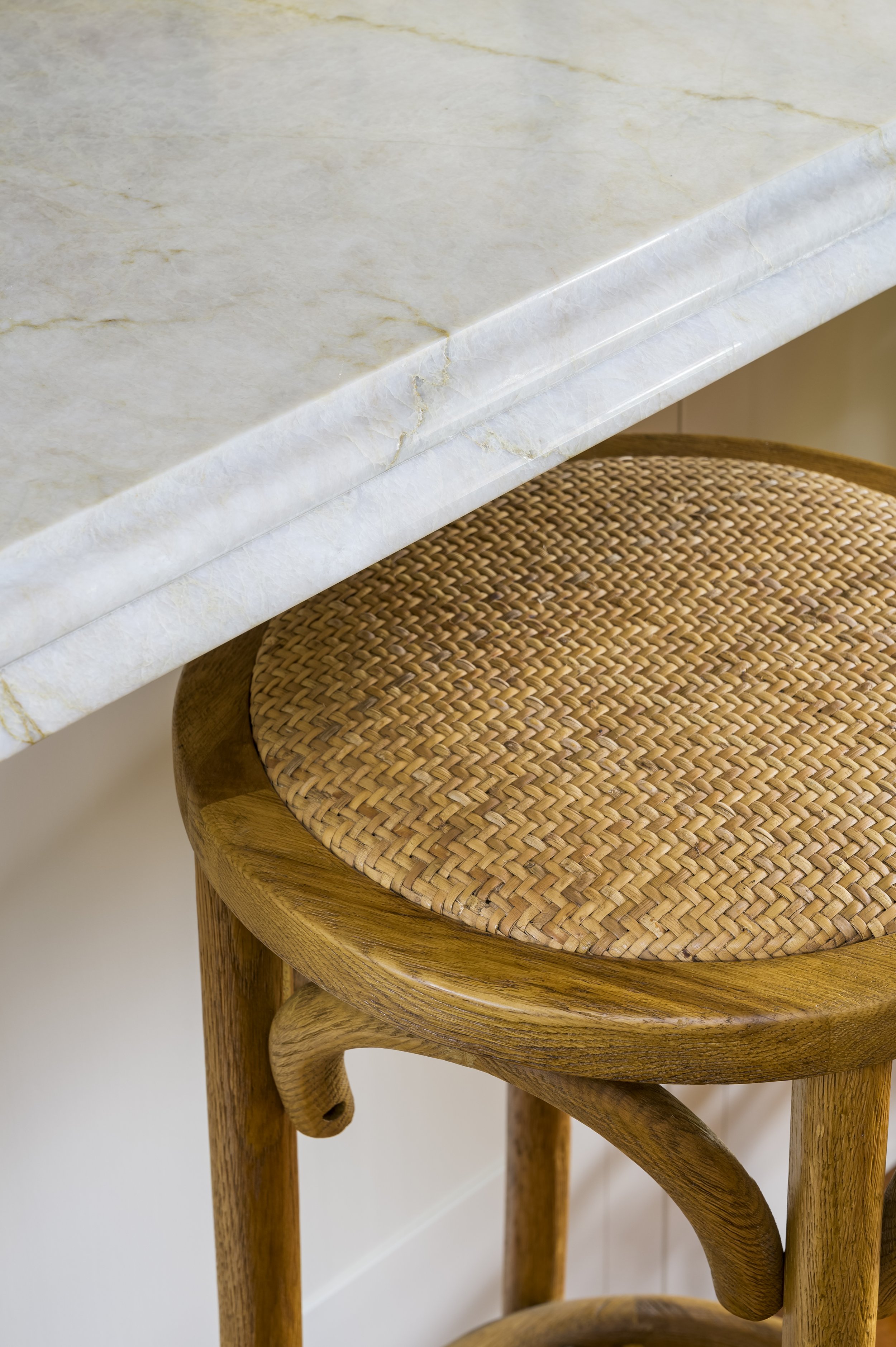 Close-up of a wooden stool with a woven rattan seat, placed under a marble countertop.