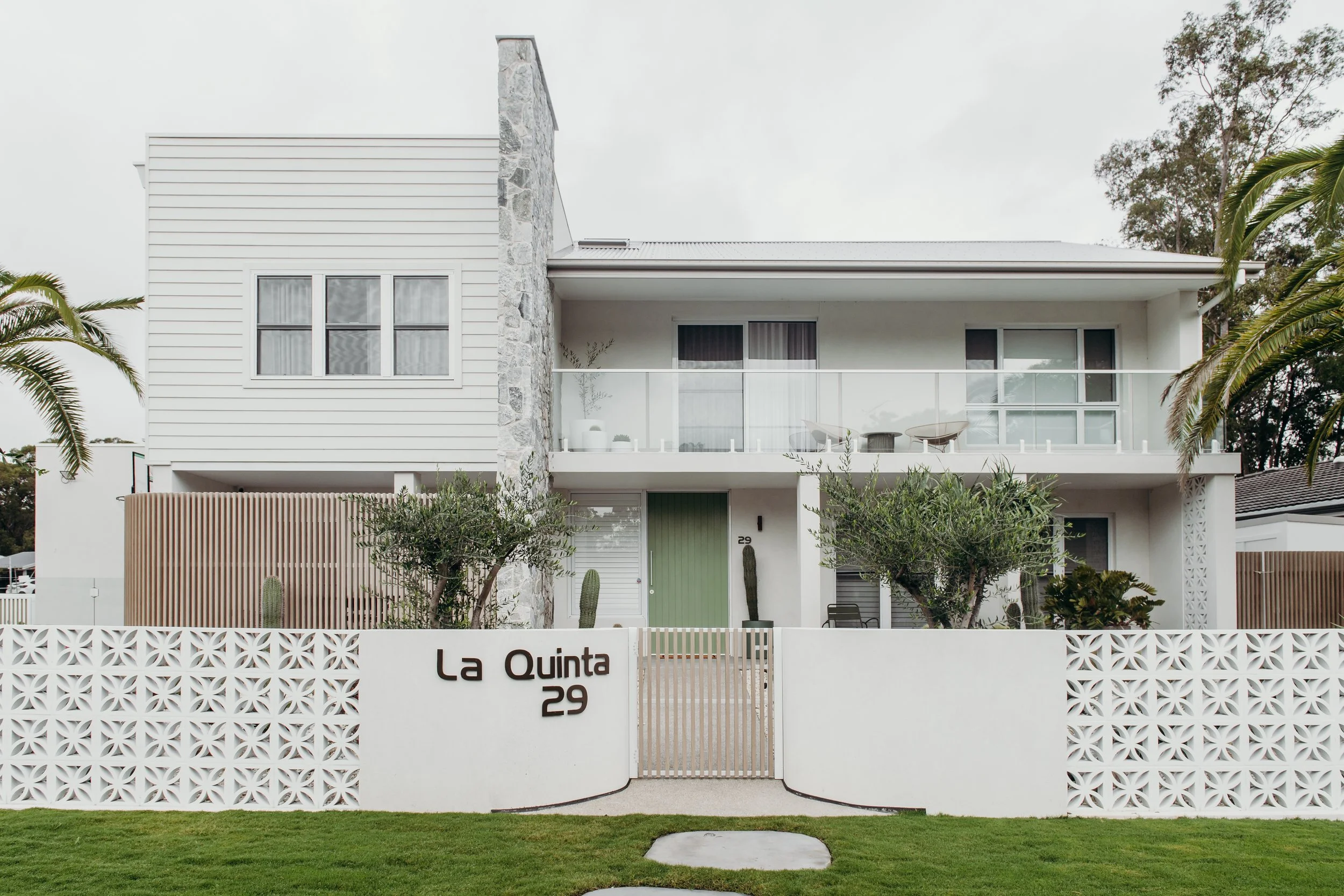 Modern two-story white house with a curved white fence, trees, and a green lawn in front. The house has large windows, a balcony with glass railing, and a stone accent wall.