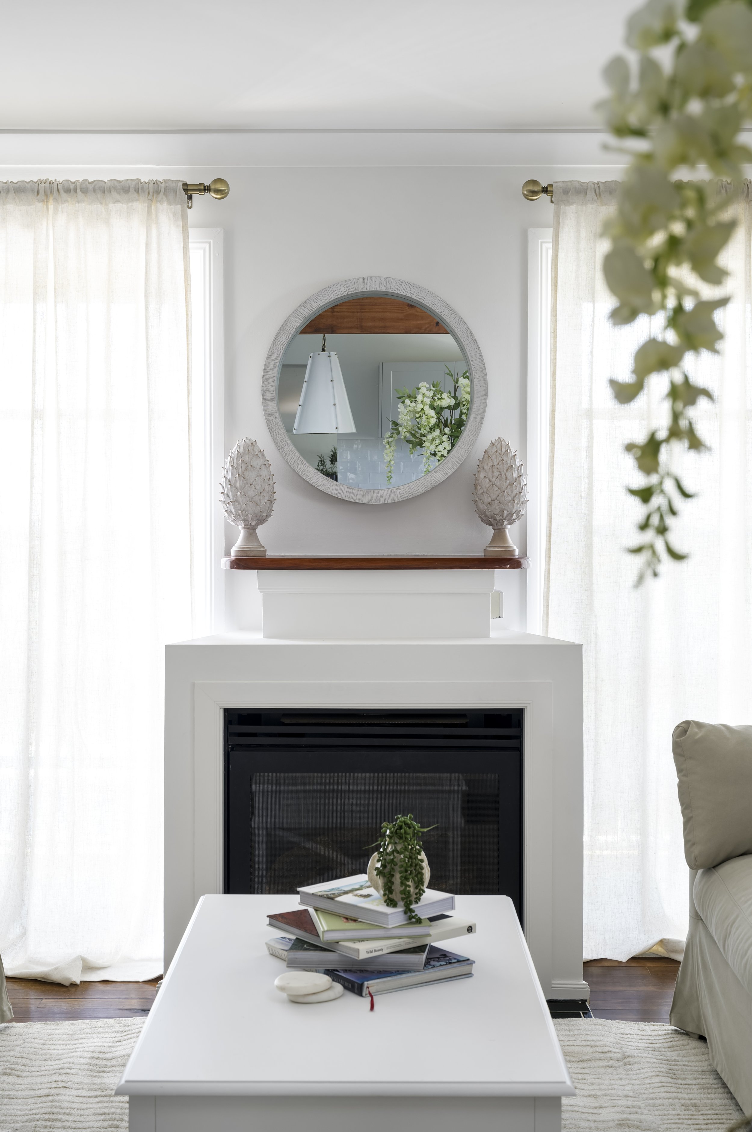 Living room with fireplace, round mirror above mantel, decorative pine cone sculptures, coffee table with books and small plant, sheer curtains, and light-colored furniture.