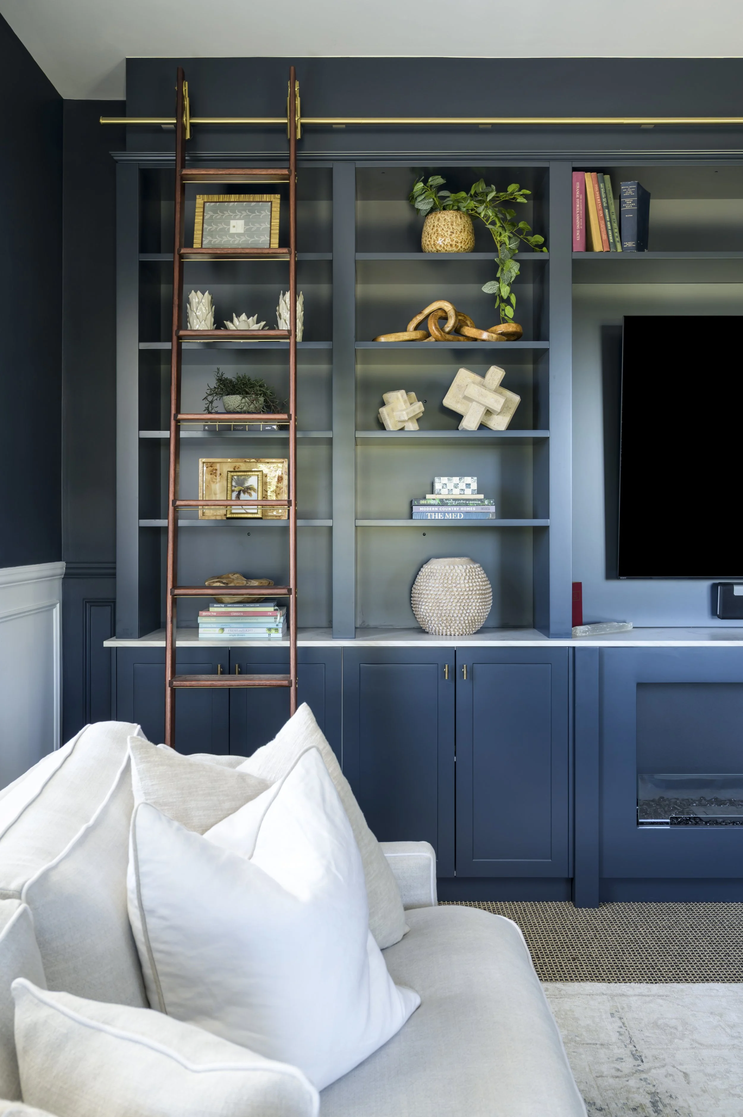 Living room with a dark blue built-in bookshelf and cabinet, filled with decorative items, books, and a potted plant, with a corner of a cream sofa with white cushions visible in the foreground.