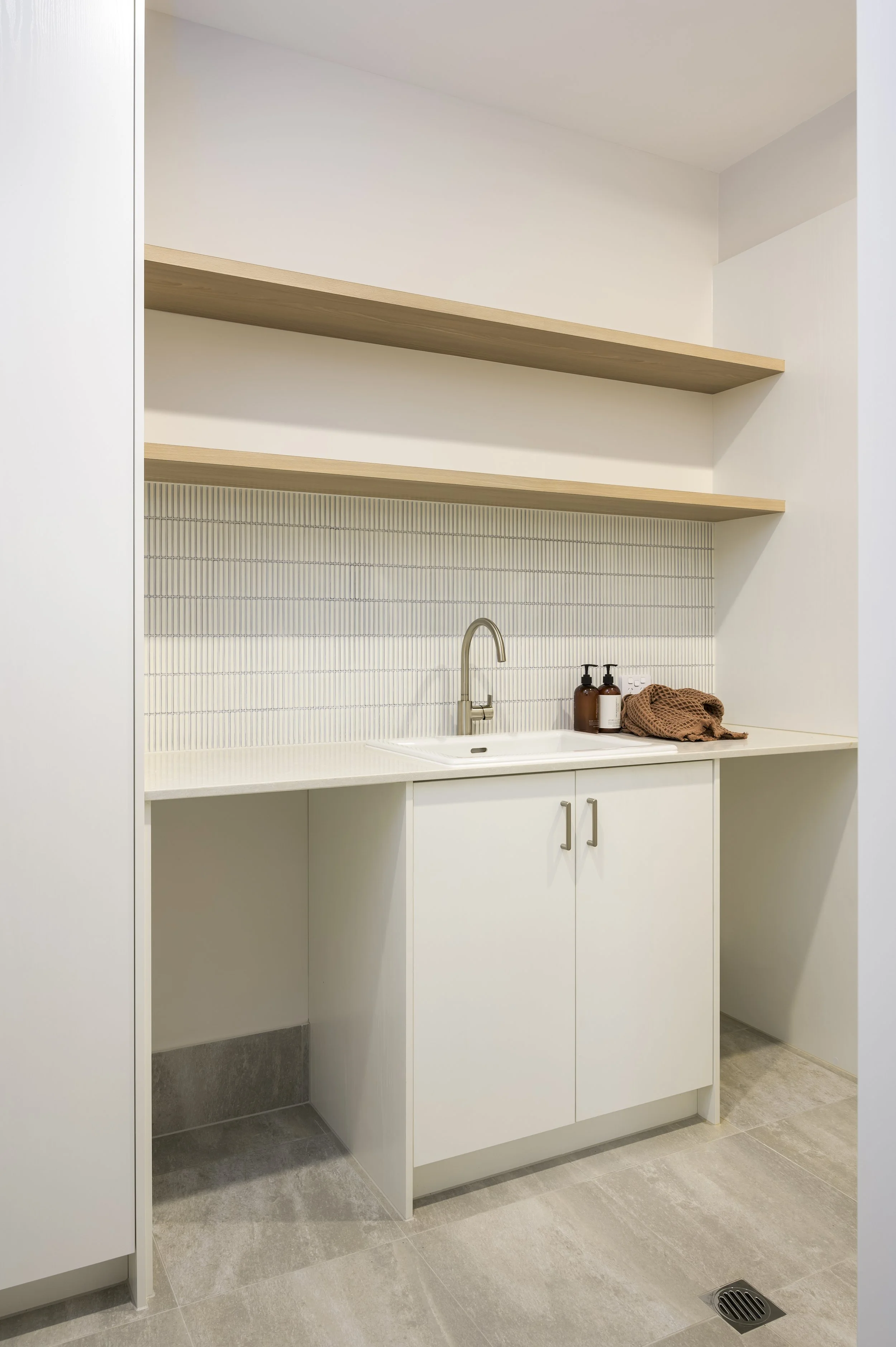 A minimalist laundry room with white cabinetry, a countertop with brown soap dispensers, a hand towel, and an open shelf above.