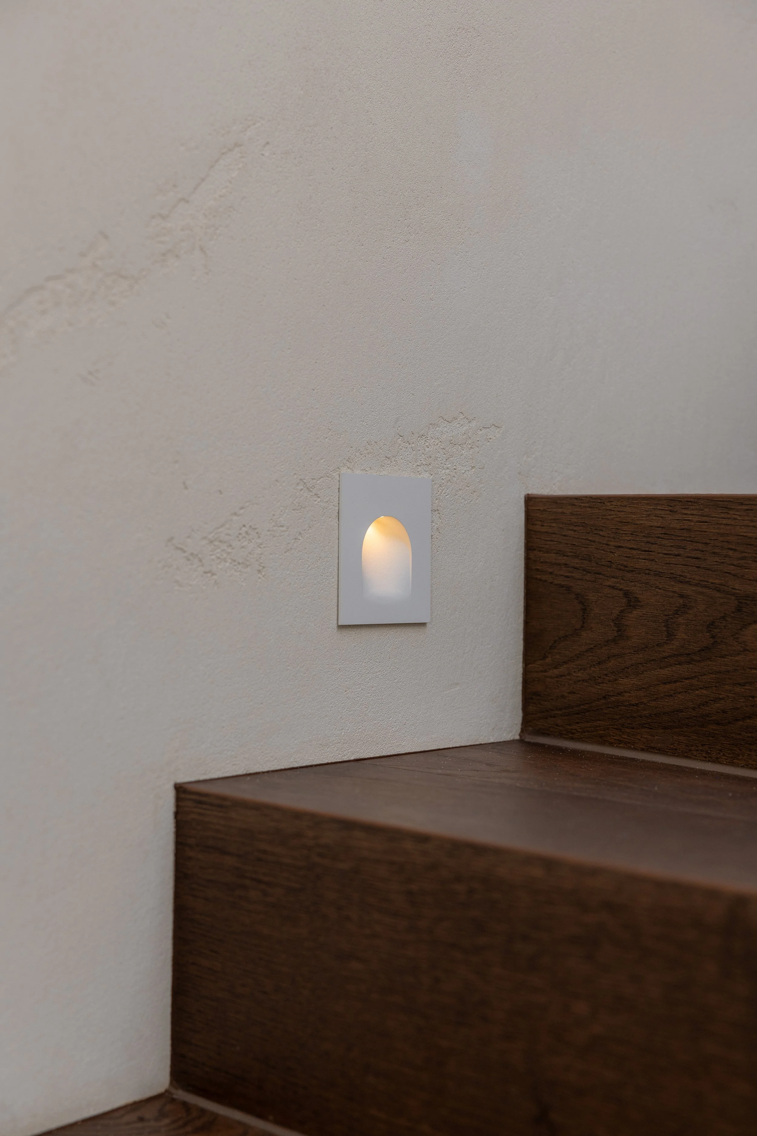 Close-up of a wall-mounted light fixture illuminating a textured, off-white wall near dark wooden staircase.