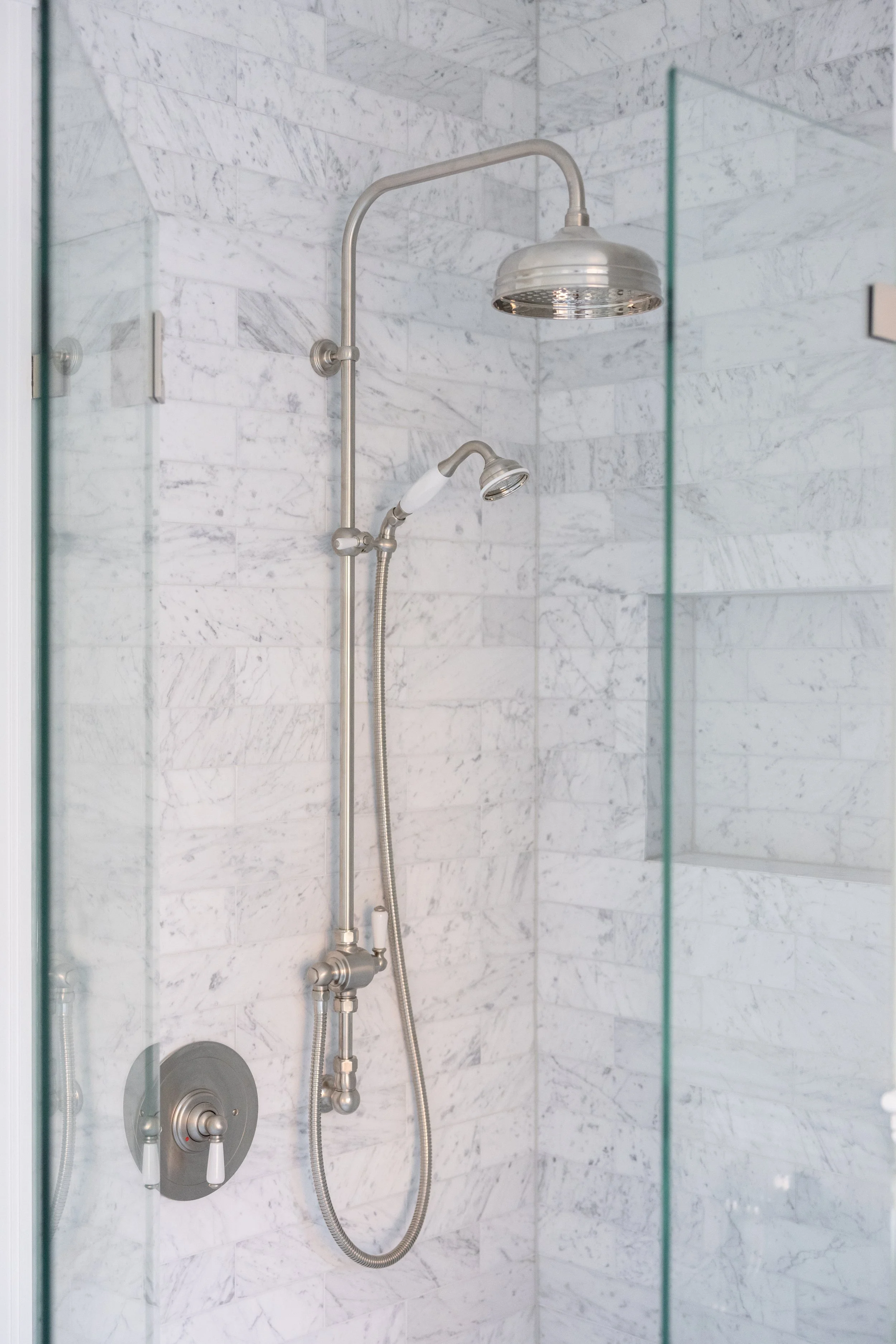 Shower with a chrome showerhead and a hand-held shower attached to a wall, set in a marble bathroom with a glass door.
