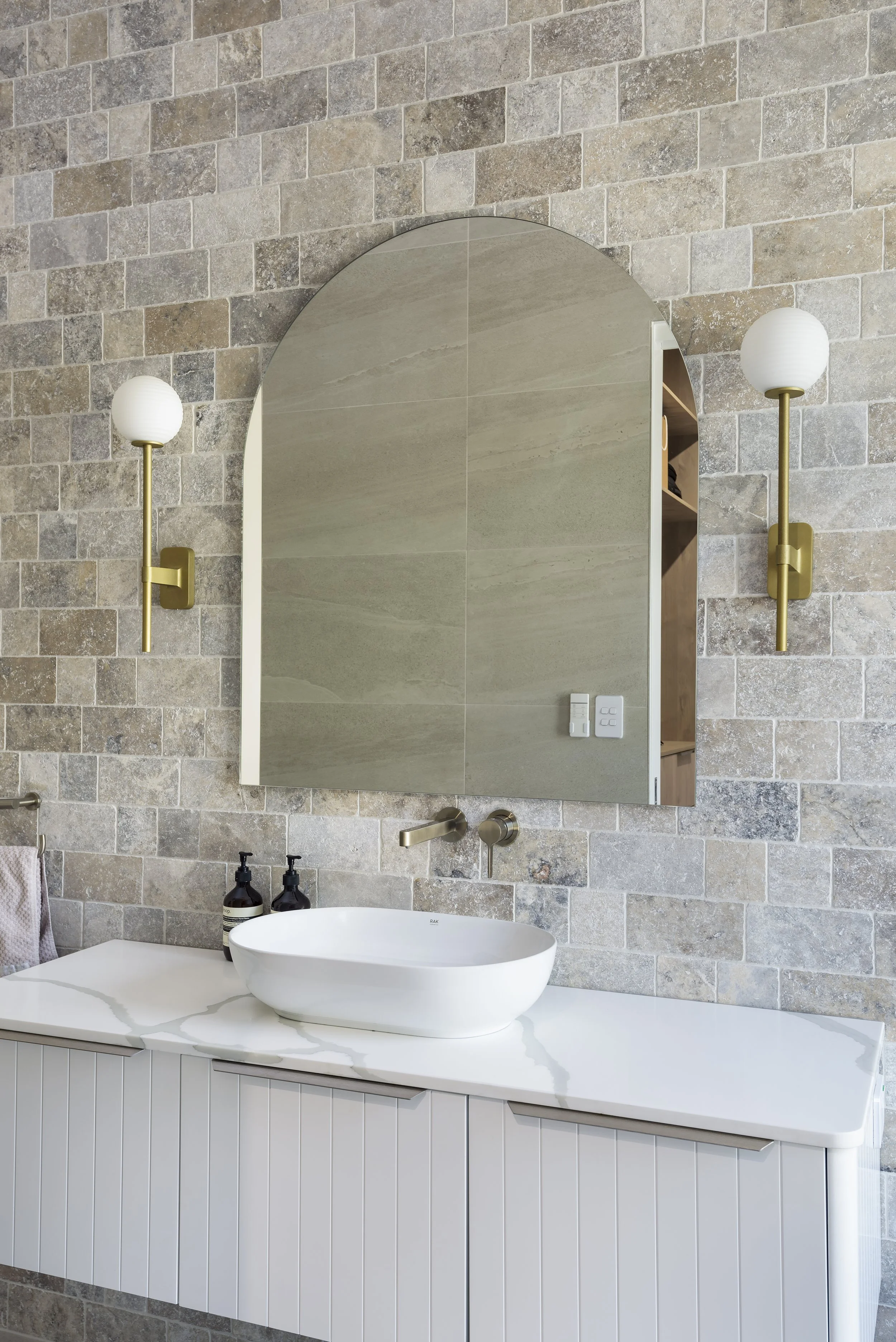 Modern bathroom with stone wall, large mirror, white vessel sink, brass fixtures, and wall-mounted sconces with brass bases and white globes.