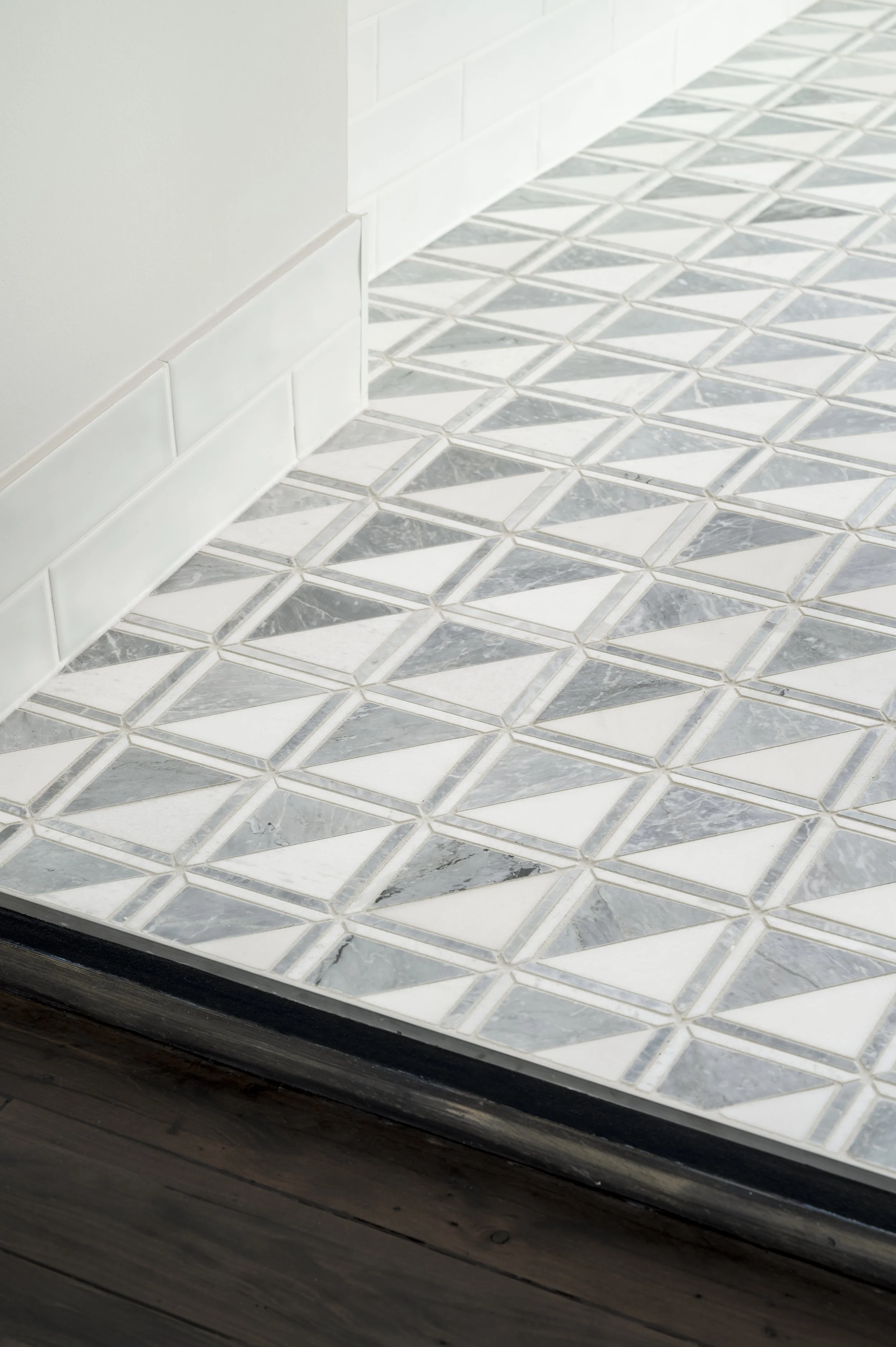 Close-up of a tiled floor with a geometric pattern in shades of white and gray near a white wall and dark wooden threshold.