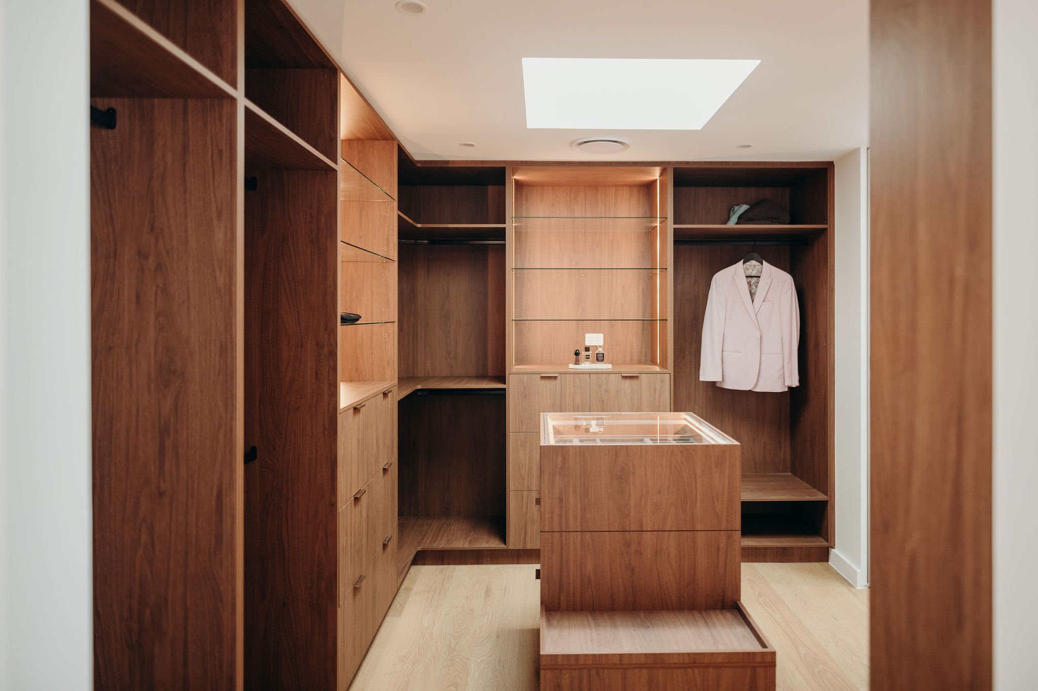 A walk-in closet with wooden shelves and drawers, a white blazer hanging on a hook, and a skylight overhead illuminating the room.