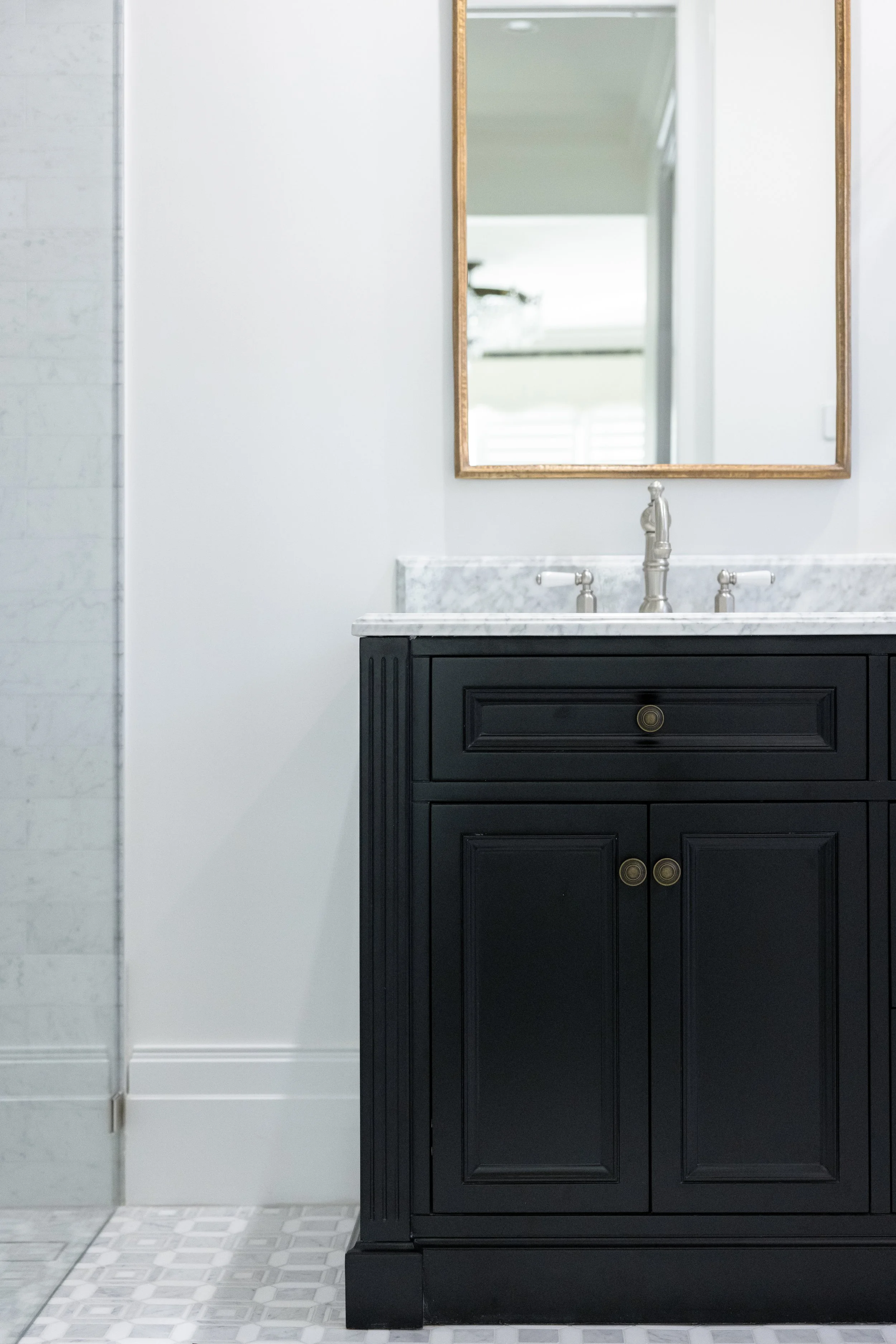 A black bathroom vanity with a marble countertop, two cabinet doors, and brass knobs, with a mirror above it and part of a white wall behind.