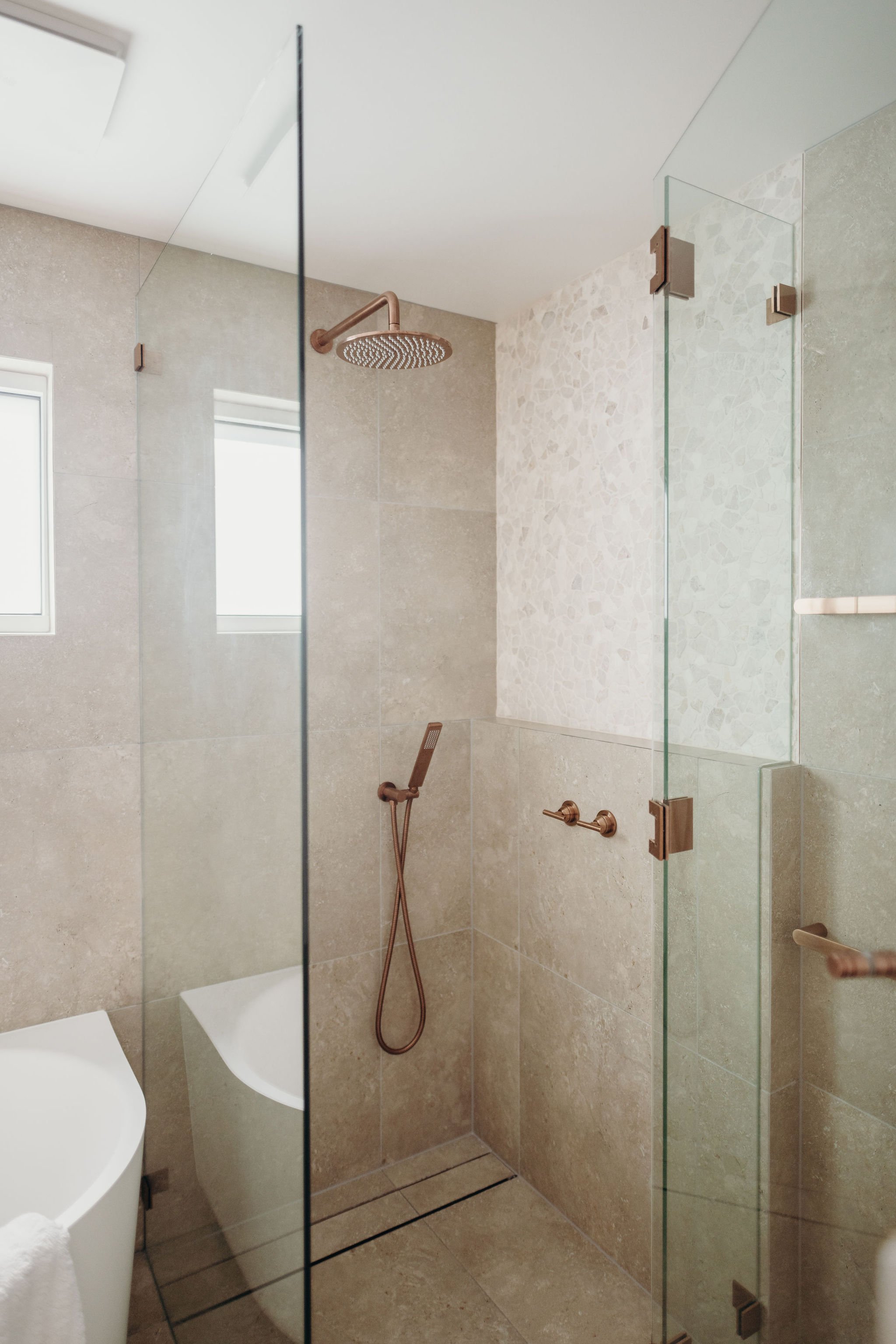 A modern walk-in shower with beige tiles and a glass enclosure. It features a round showerhead and a handheld shower wand with a bronze finish. The shower has a small window allowing natural light to enter.