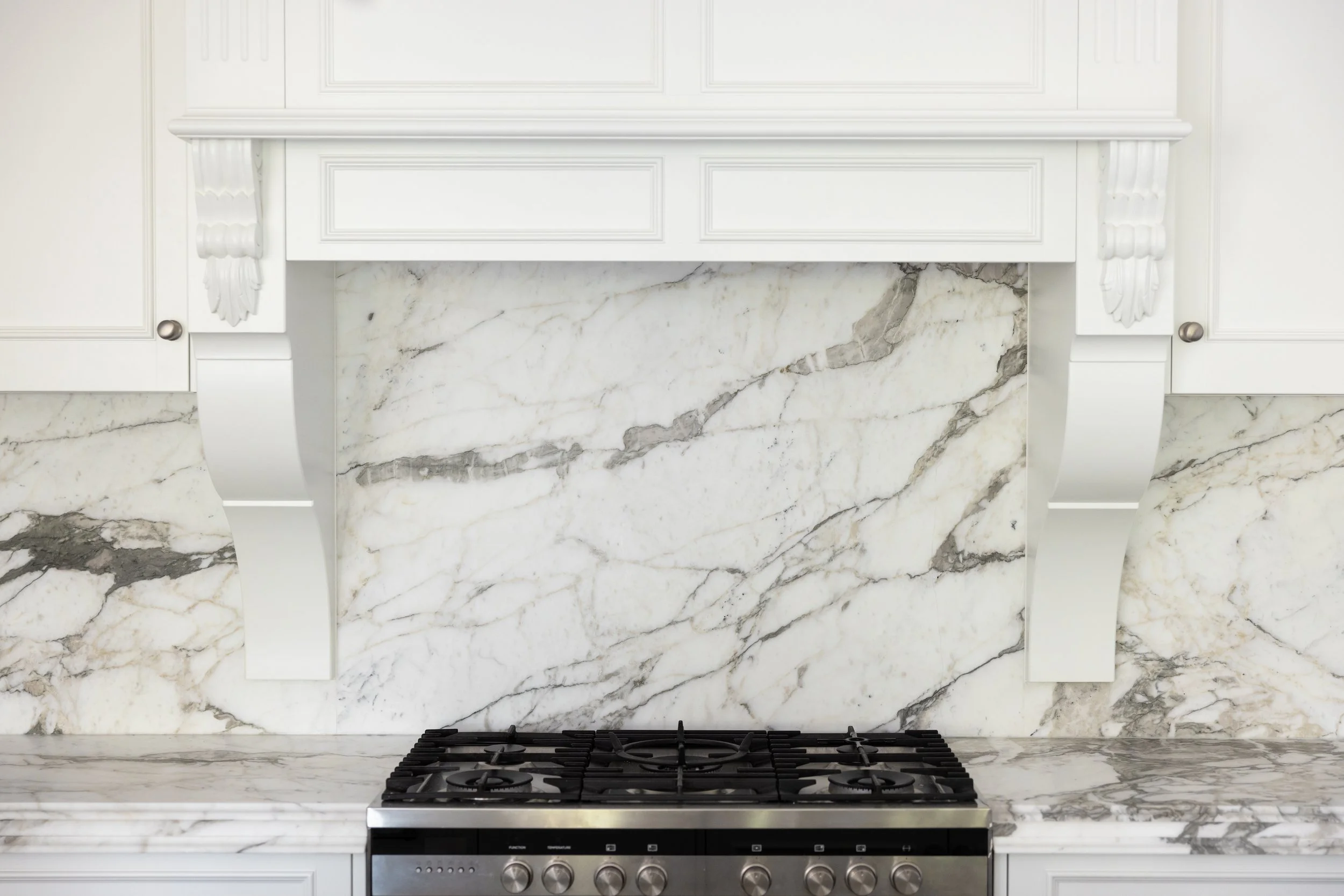 Close-up of a modern kitchen featuring a white marble backsplash with gray veins, white cabinetry with decorative molding, and a stainless steel gas cooktop.